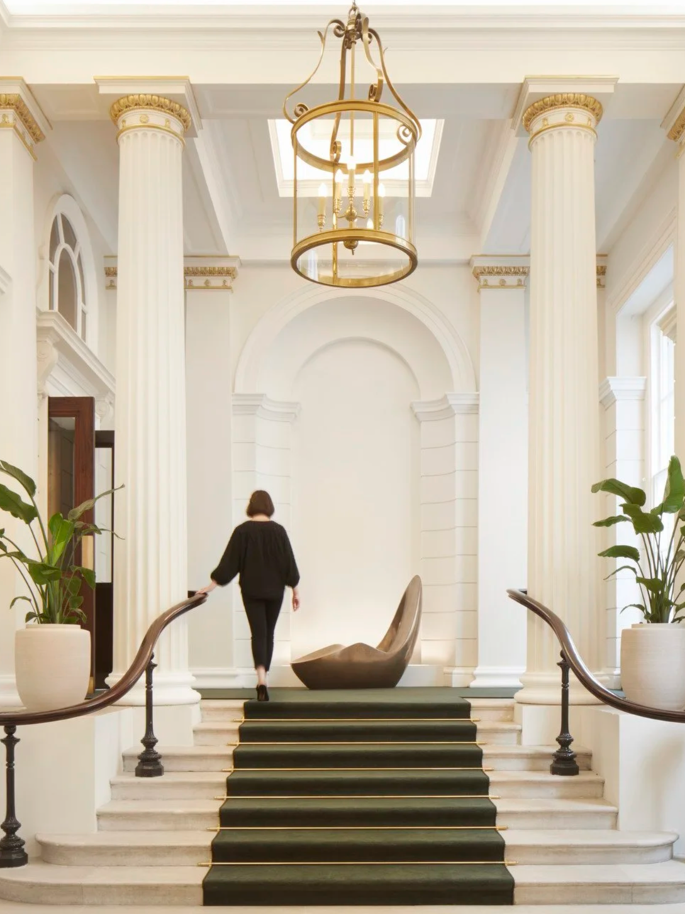 Elegant hotel lobby entrance with grand staircase, large potted plants, tall white columns with gold accents, a chandelier, a modern sculpture, and a woman walking up the stairs.