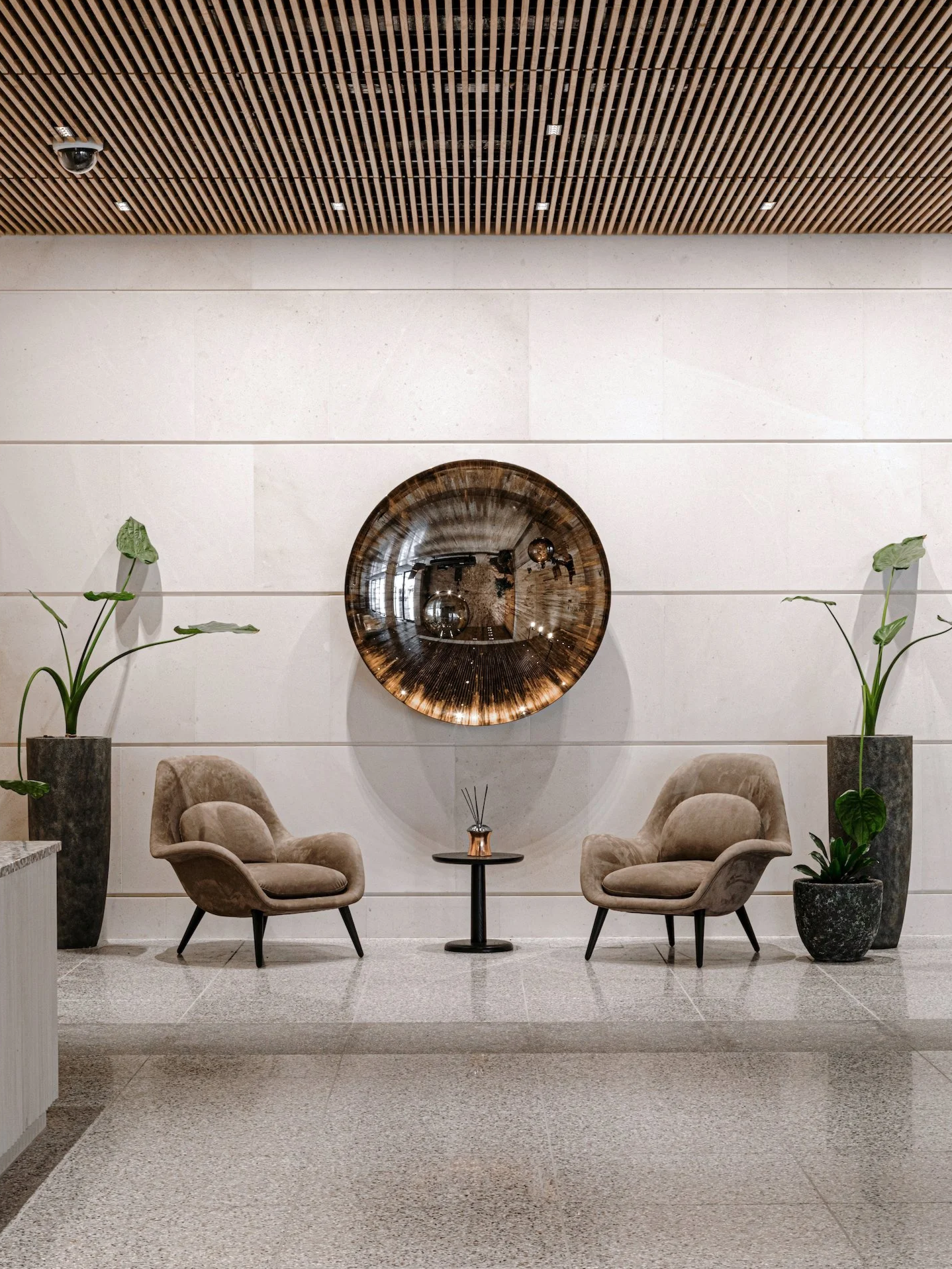 Modern lobby with two beige armchairs facing each other, a small black table with reed diffusers between them, two tall potted plants, a large round mirror on the wall, and a wooden slatted ceiling.