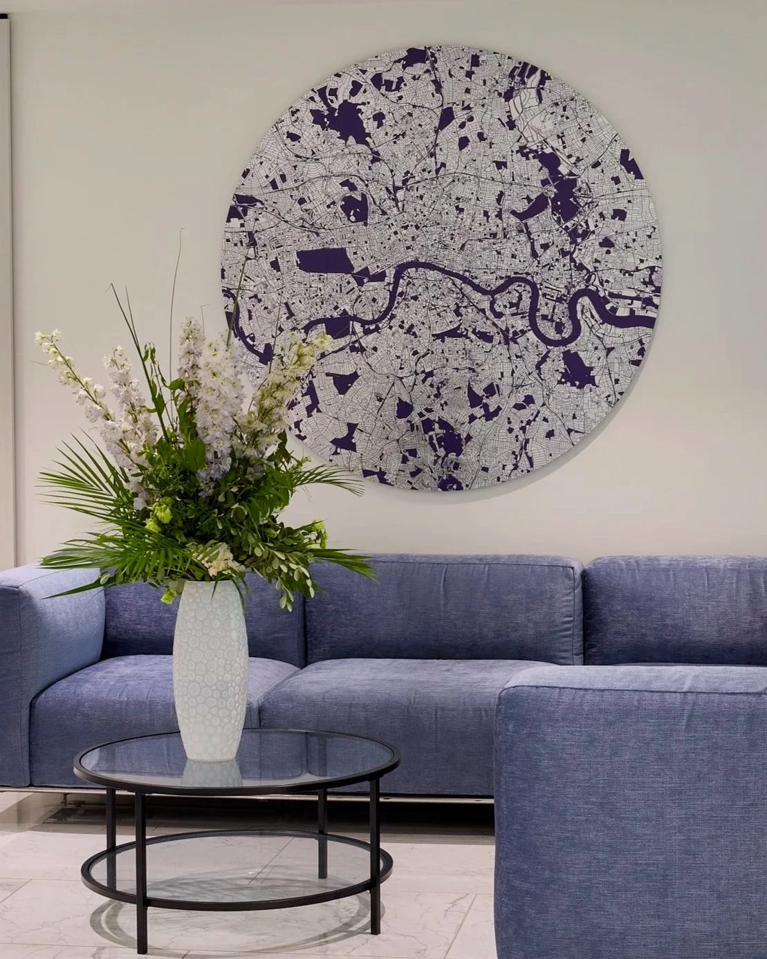 A blue sofa with a circular black metal and glass coffee table in front of it. A tall white vase with a floral arrangement of white and green flowers, including long leaves, is on the table. On the wall behind, there is a large round map of a city wi