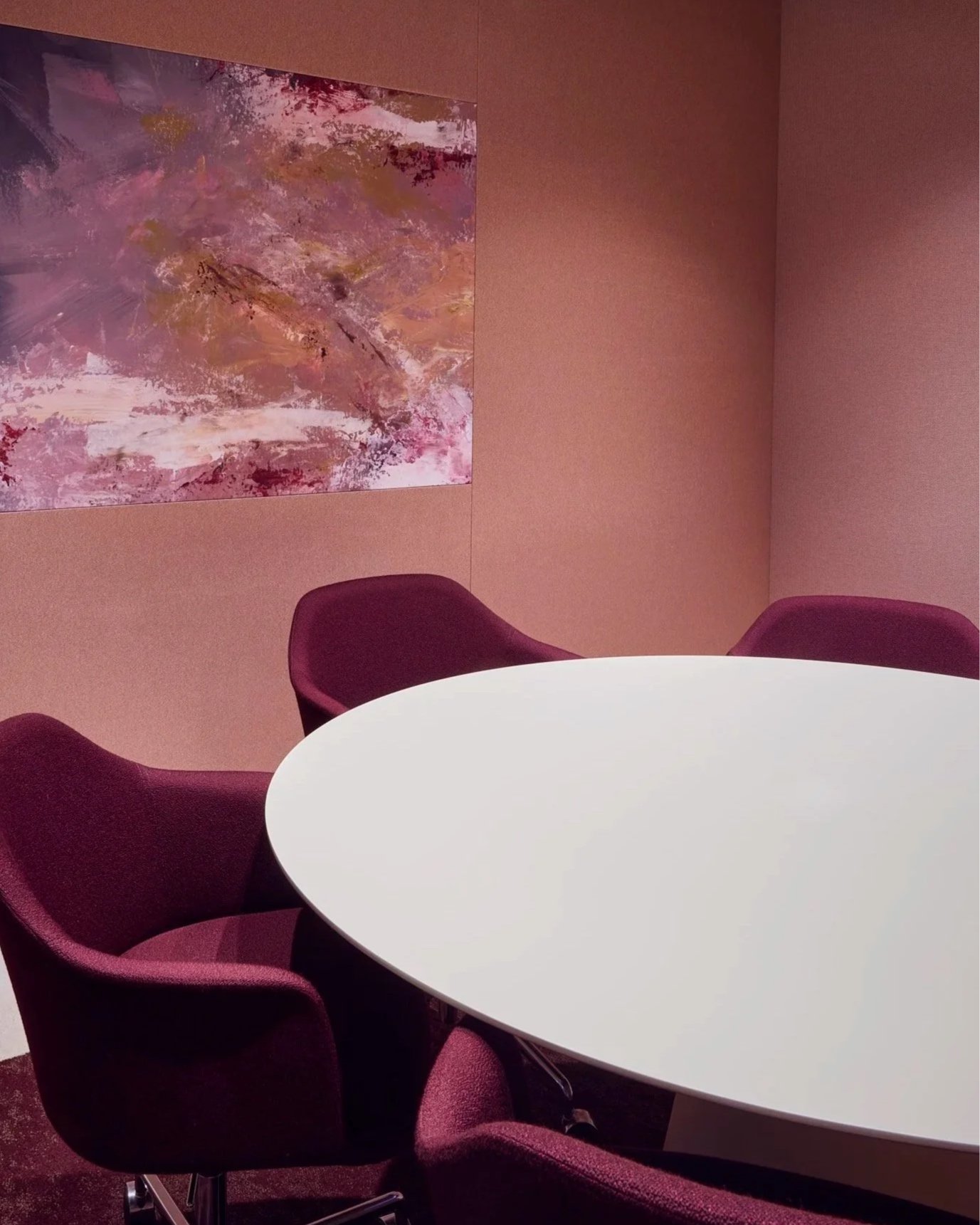 A round white table surrounded by burgundy chairs in a room with pink walls. There is an abstract pink and purple painting on the wall.