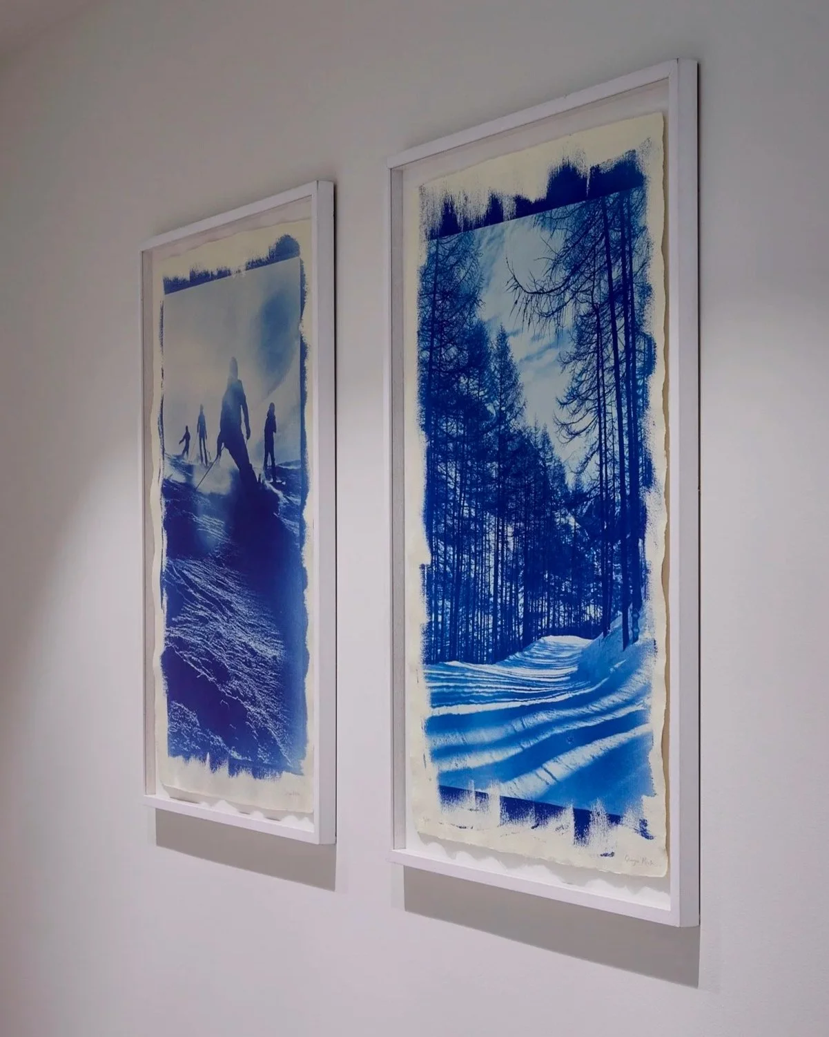 Two framed pictures of snowy winter forests hanging on a white wall.