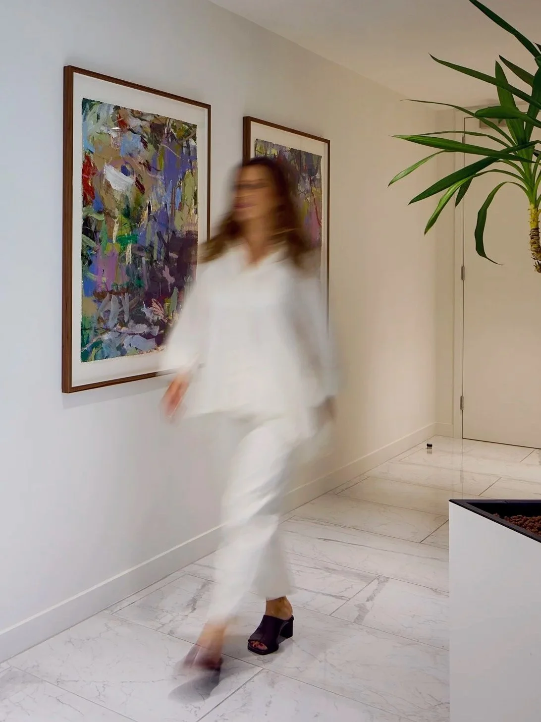 A woman dressed in white with black high-heeled shoes walking past colorful abstract paintings on a white wall in an art gallery or modern interior space, with a TV or speaker on the floor in the corner and a large green plant on the right.