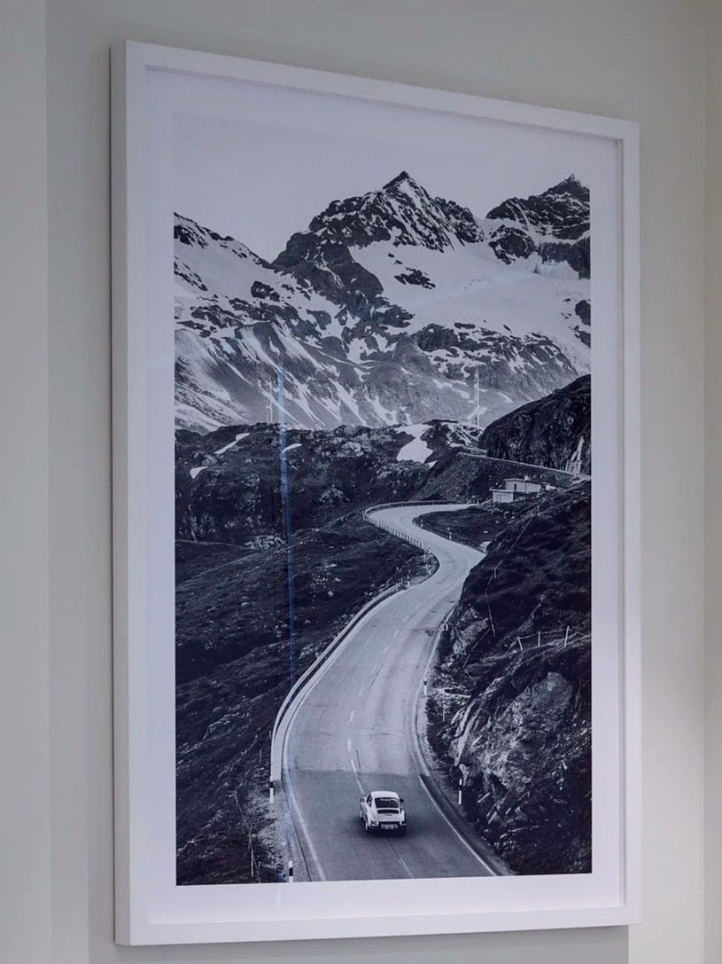 Black and white photograph of a winding mountain road with a white car driving on it, surrounded by rocky terrain and snow-capped mountains in the background.