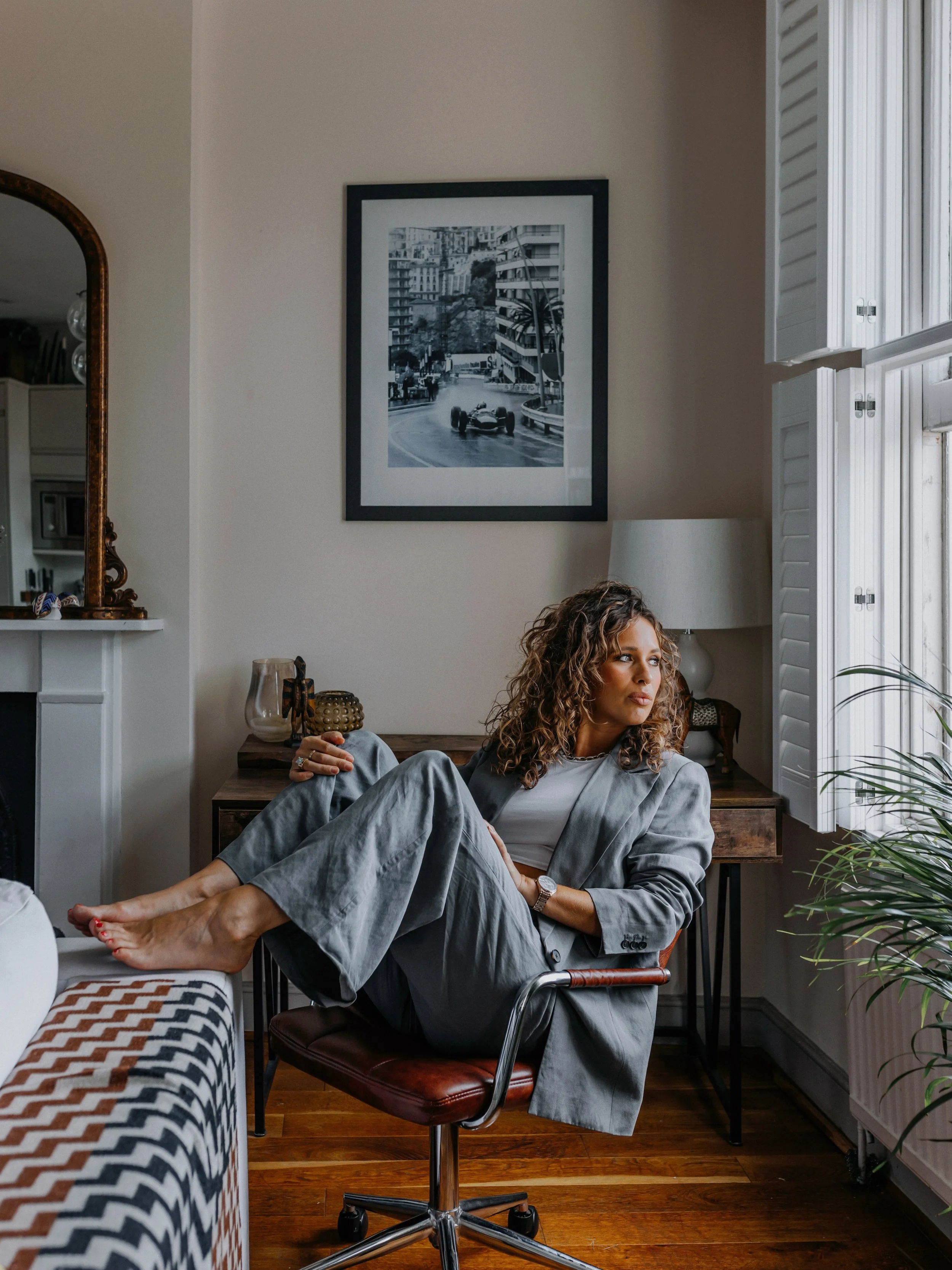 A woman with curly hair in a gray blazer and pants sitting on a rolling chair with her feet resting on a table in a living room, looking to the side. The room has a framed black and white photo of a city scene on the wall.