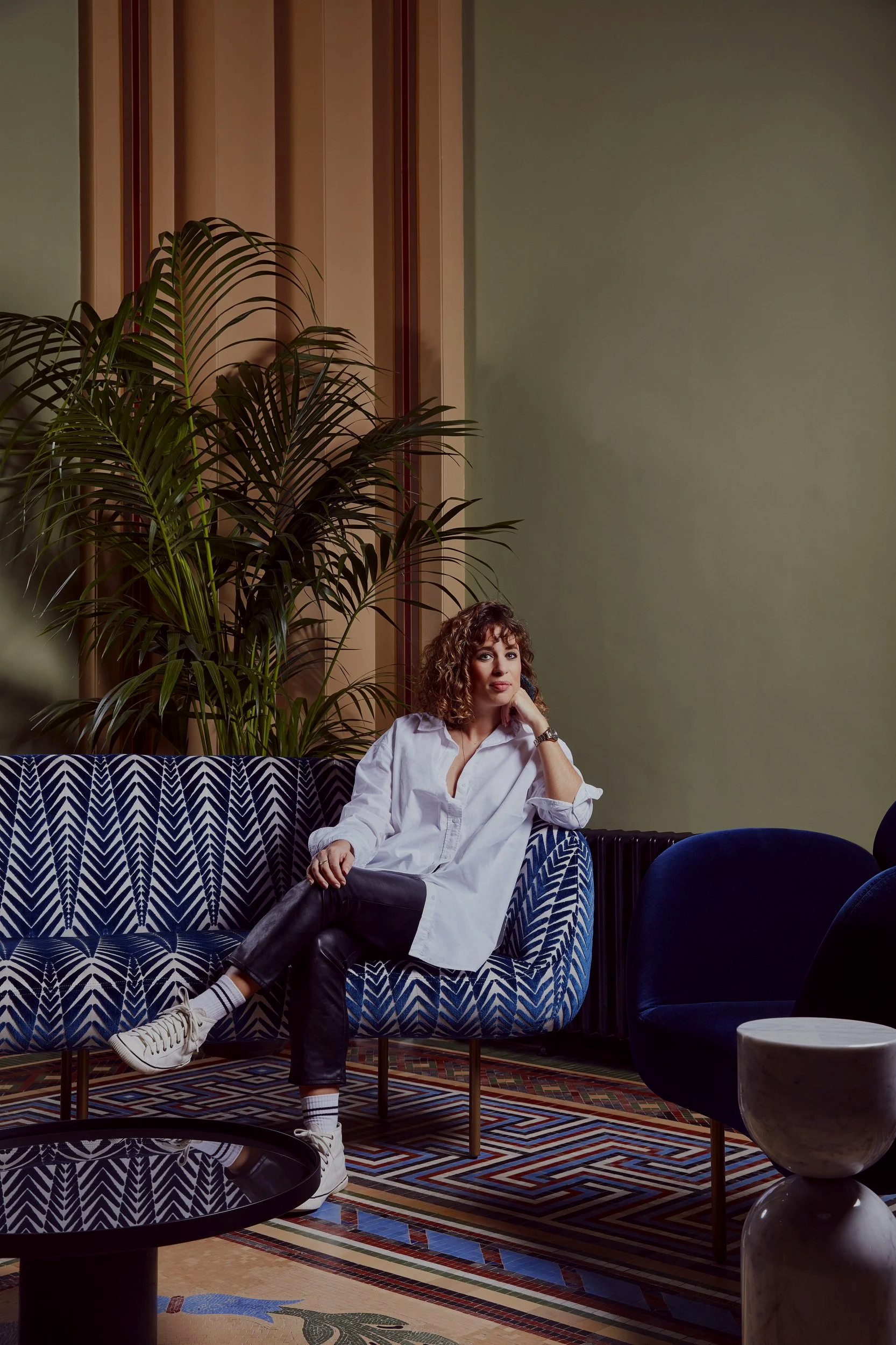Woman sitting on a patterned blue and white sofa with one leg crossed over the other, wearing a white shirt, black pants, white sneakers, and striped socks, in a modern interior with a green wall, wooden paneling, a large leafy plant, and various furniture including a round marble-topped table and a dark blue chair.