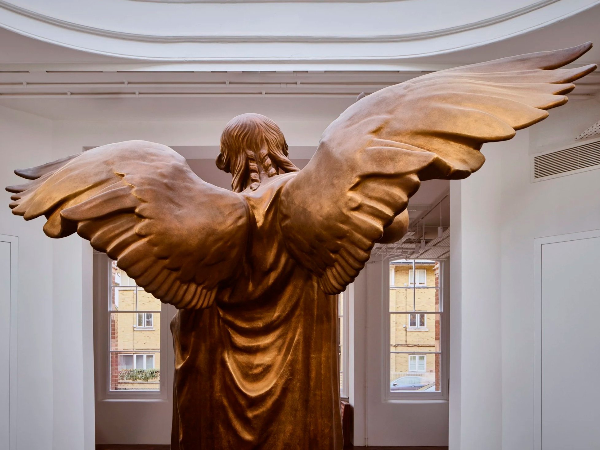 A large wooden sculpture of an angel with outstretched wings, facing away from the camera, inside a bright room with windows showing buildings outside.