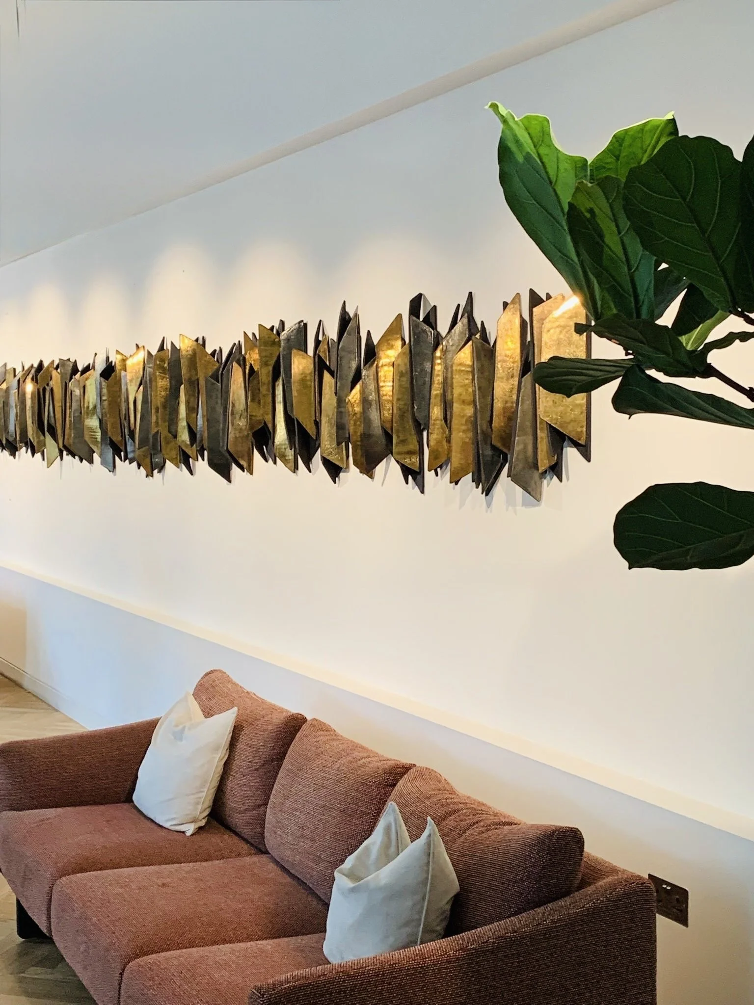 A reddish-brown upholstered couch with multiple white pillows, positioned against a white wall. A large green leafy plant is partially visible on the right side, and a long, decorative, metallic gold and black wall hanging is mounted above the couch.