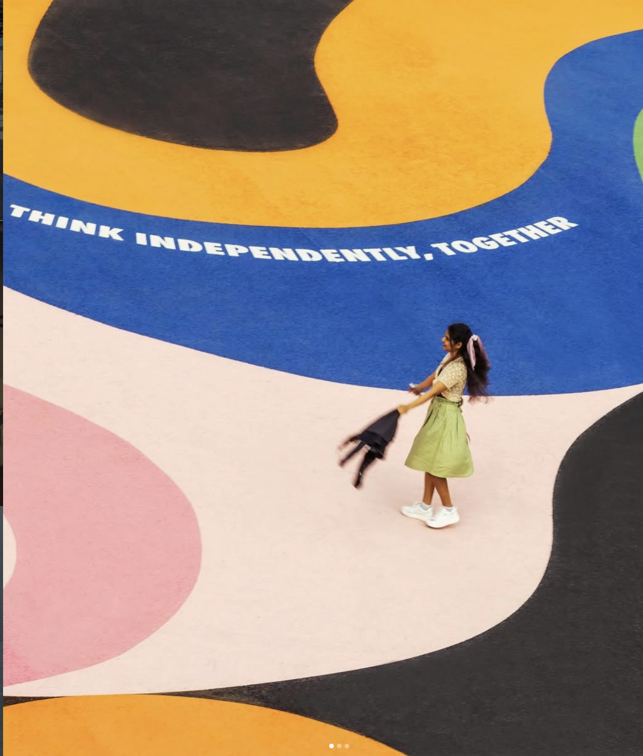A girl in a yellow skirt and white sneakers is holding a black jacket, standing on a colorful mural floor with the phrase "Think independently, together" written on a blue section.