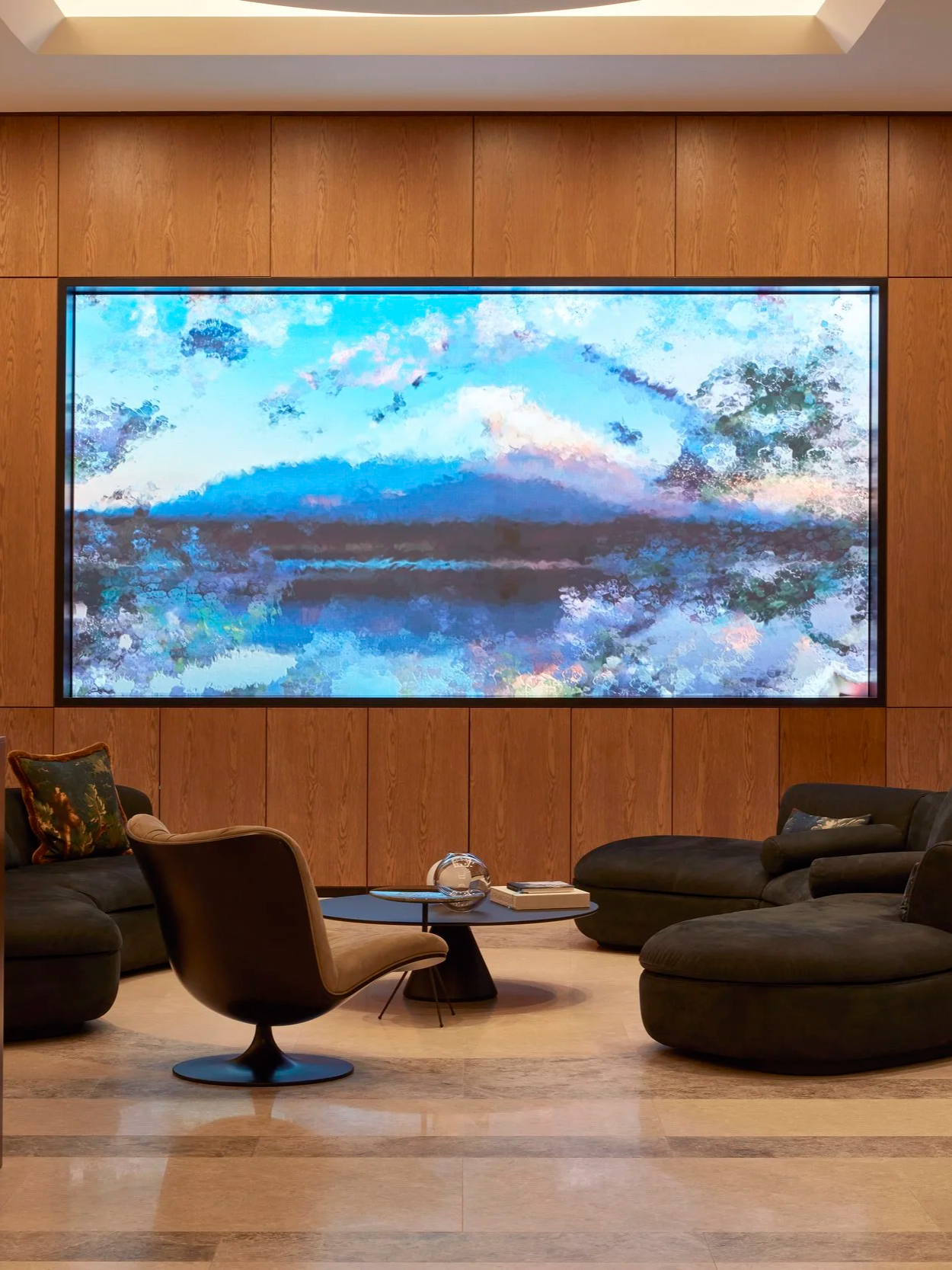 Modern living room with dark sofas, a black chair, and a round coffee table with a glass orb and books. A large digital artwork of a mountain landscape with blue, white, and pink colors is mounted on a wood-paneled wall, illuminated by ceiling lighting.
