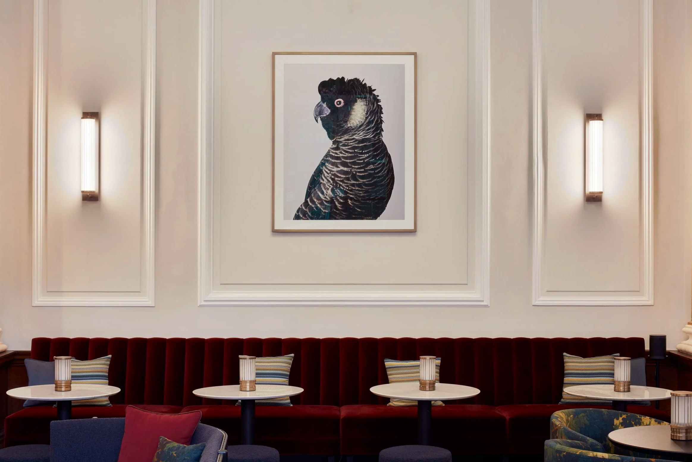 Interior dining area with a large framed bird artwork on the wall, red upholstered seating, round tables, and blue and patterned chairs.