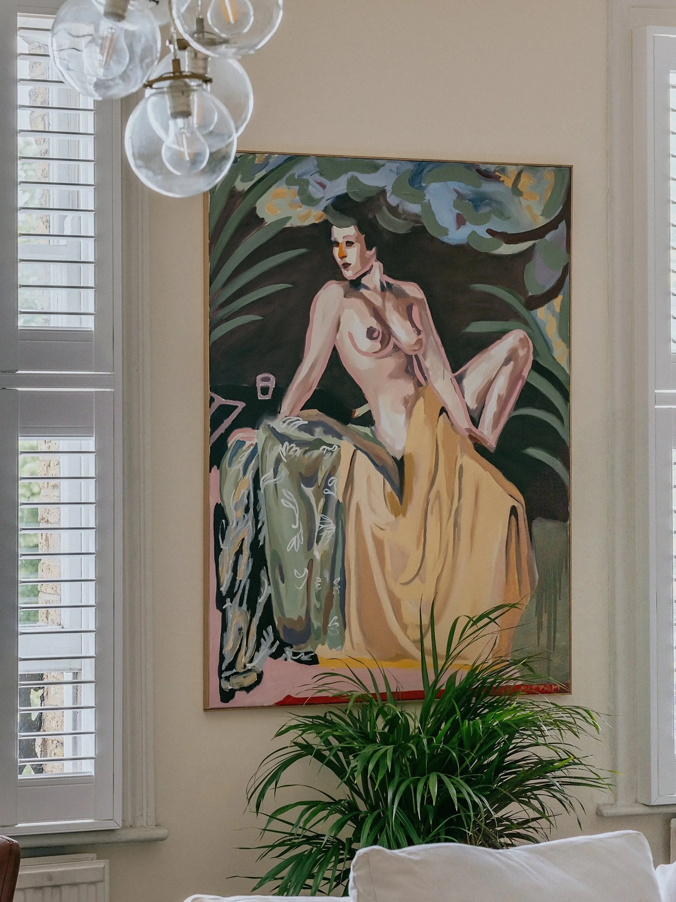 A colorful painting of a nude woman with dark hair sitting among large green leaves, hung on a light-colored wall with shutters on the window nearby, and a potted plant in the foreground.