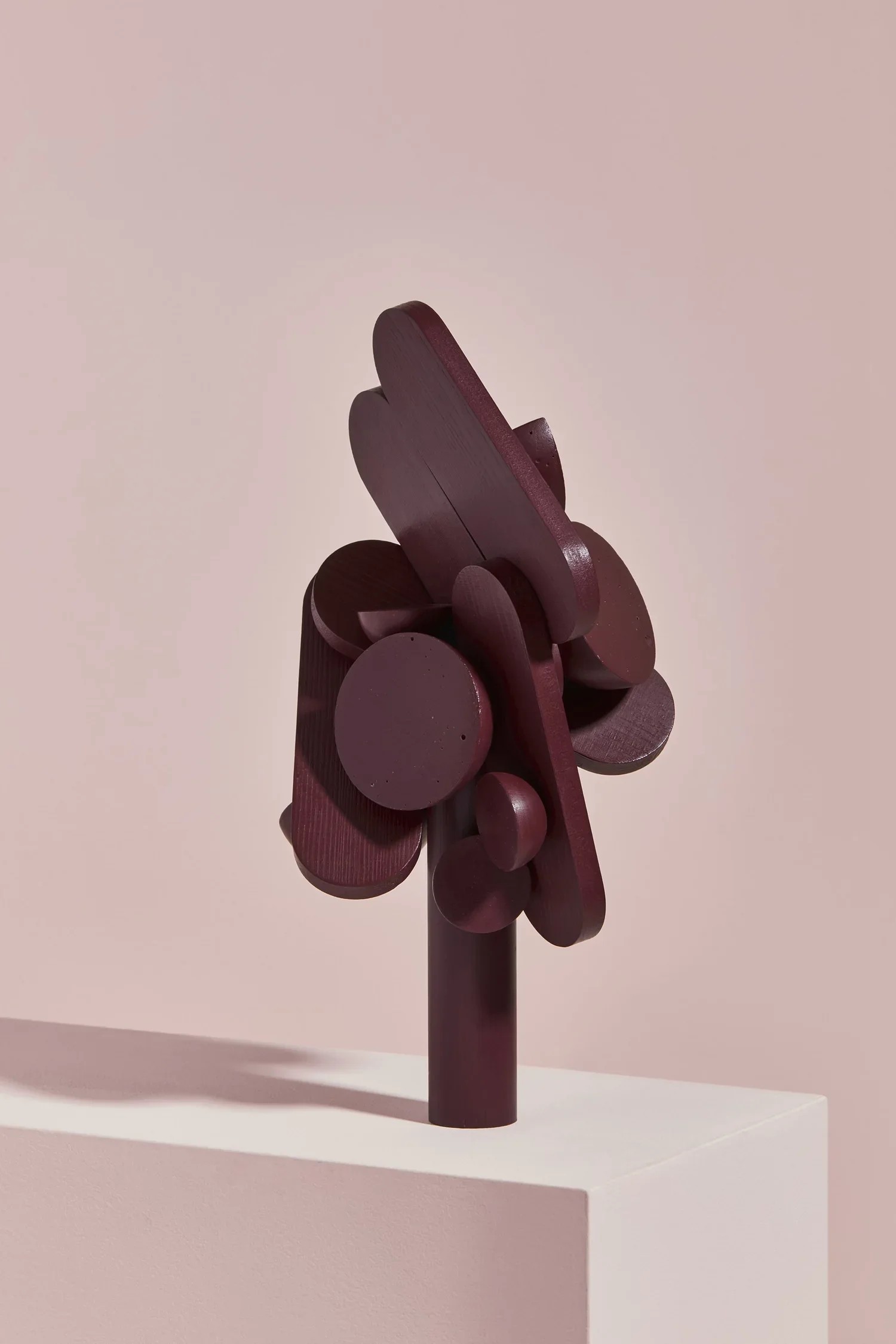 A sculpture made of dark red, rounded wooden shapes stacked and arranged to resemble a flower, mounted on a cylindrical pedestal on a white display surface against a soft pink background.