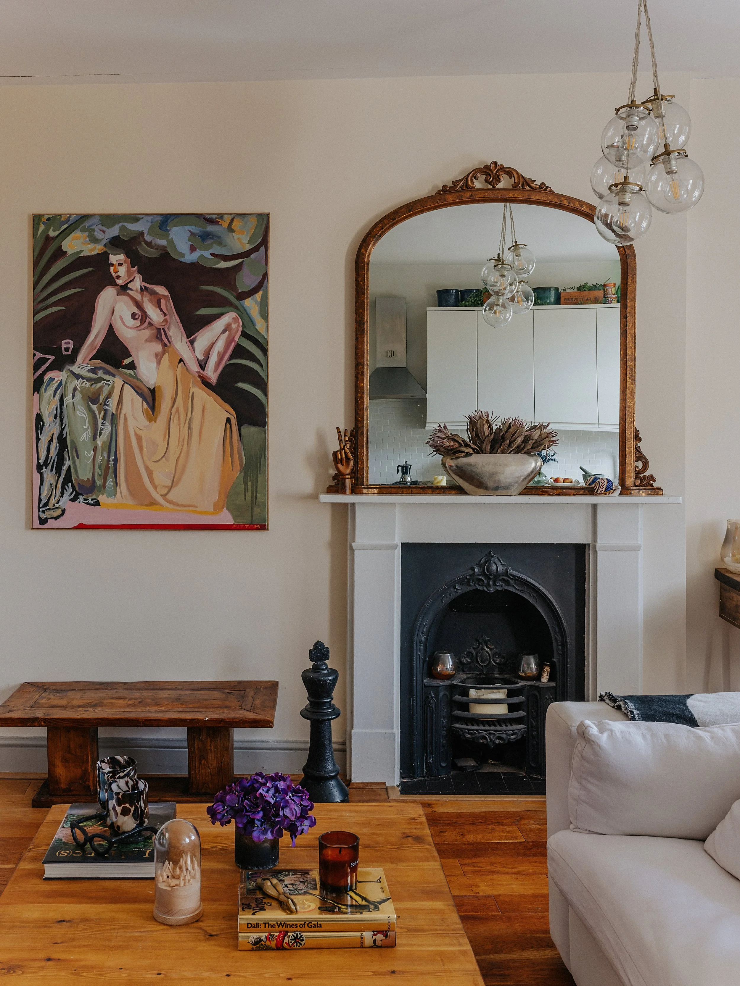 Living room with wooden coffee table, purple flowers in a black vase, books, candles, and decorative objects. A white sofa on the right, a fireplace with a mirror above, and a colorful painting of a nude woman on the left wall. Modern chandelier hang