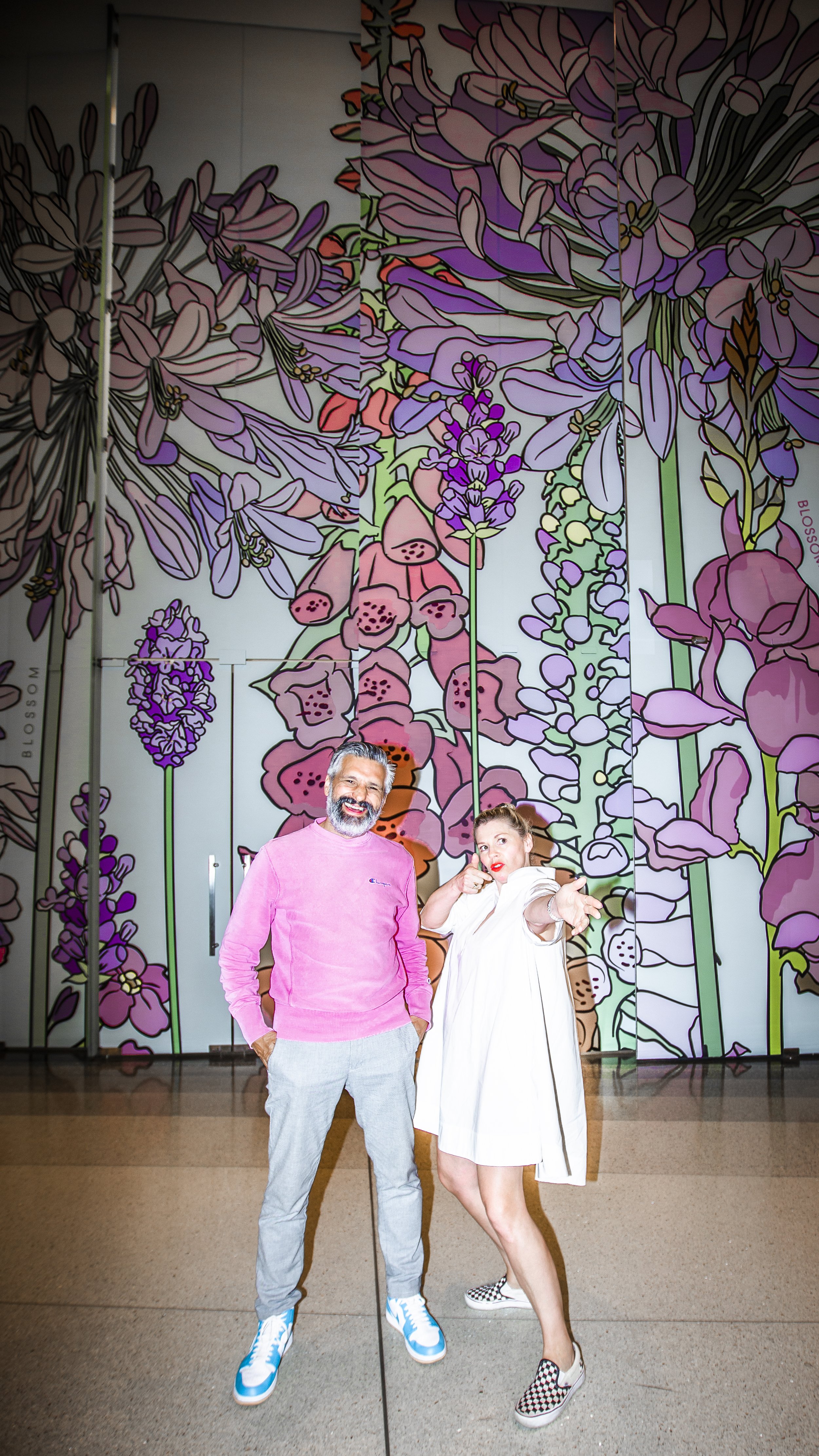 Two people, a man and a woman, posing in front of a large colorful floral wall. The man is wearing a pink sweatshirt and light gray pants, and the woman is wearing a white dress with checkered slip-on shoes. They are smiling, and the woman is holding her hand out toward the camera.