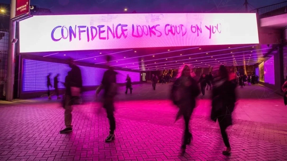 People walking on a sidewalk at night near a large digital billboard displaying the message 'Confidence looks good on you' in purple and pink lights.