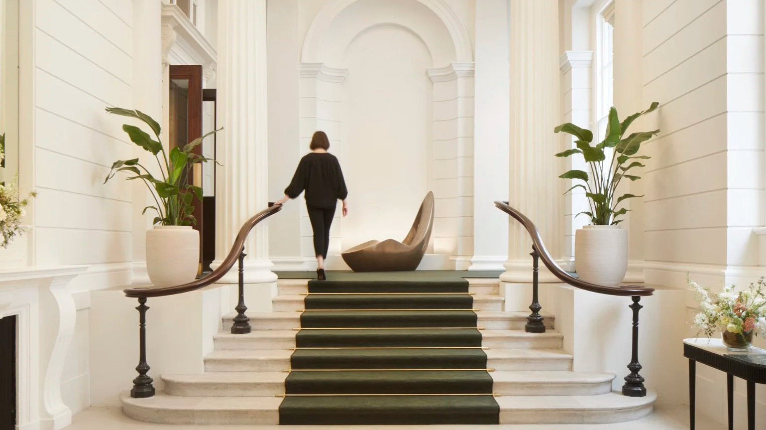 Interior of a luxurious building with white walls, marble staircase with a green runner, decorative plants in large white pots, and a woman walking up the stairs.
