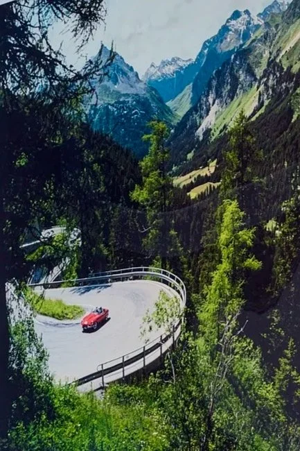 A winding mountain road with a red car driving on it, surrounded by green trees and forested slopes, with snow-capped mountains in the distance.