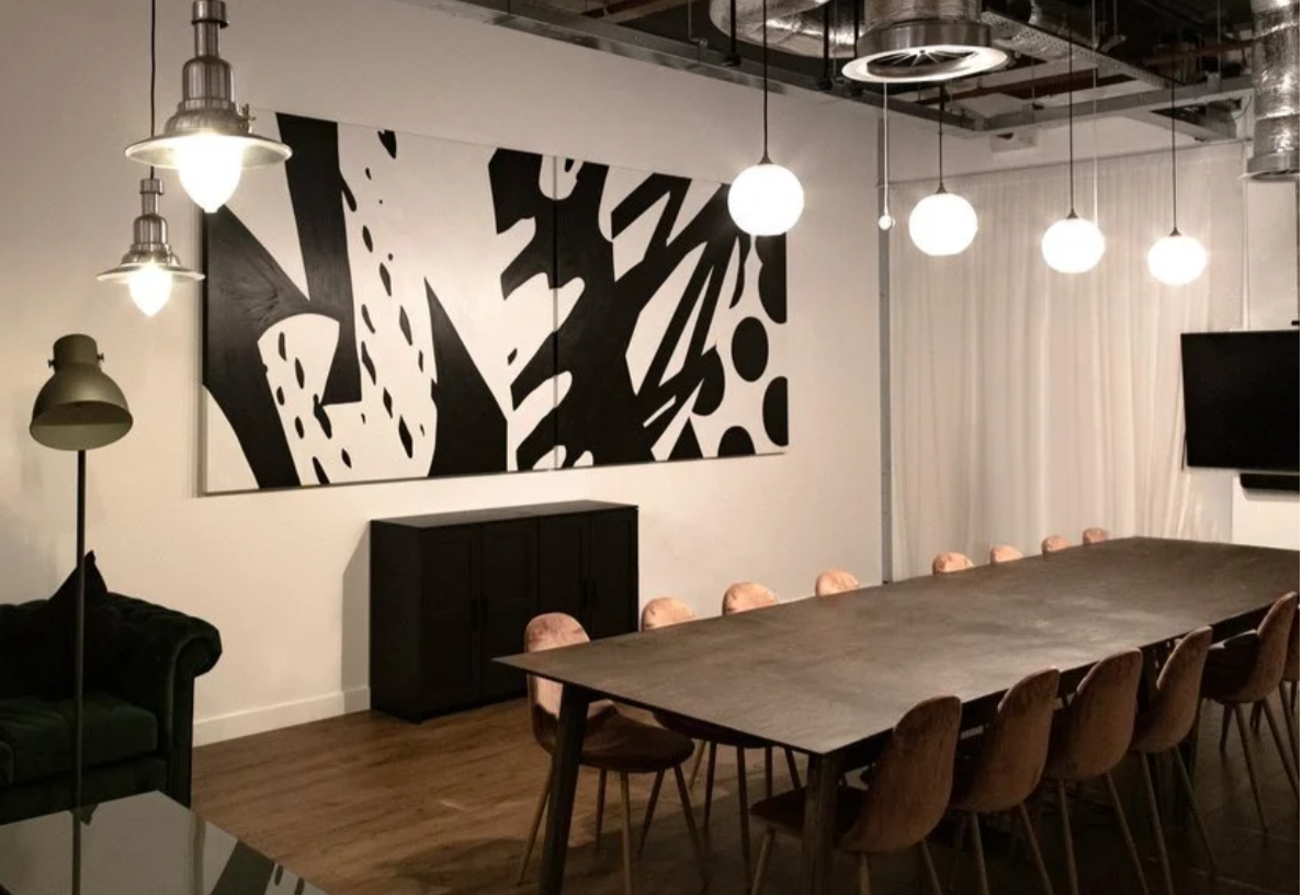 A modern conference room with a long wooden table, pink chairs, large abstract black and white artwork, hanging ceiling lights, a TV, a black cabinet, a black sofa, and a standing floor lamp.