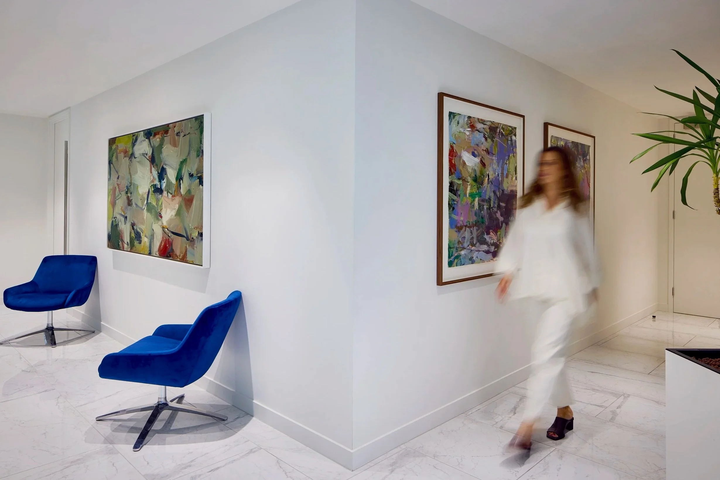 Interior of a modern, minimalist room with white walls, featuring two abstract colorful paintings, blue chairs, a woman with brown hair in a white outfit walking, and a large green plant in the corner.