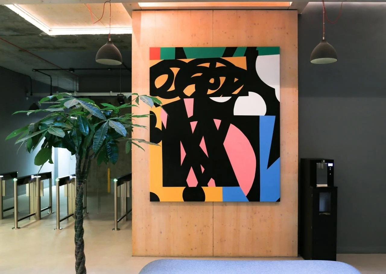 Abstract colorful artwork on a wall in a modern interior space, with a large potted plant and a water dispenser nearby, and ceiling pendant lights.