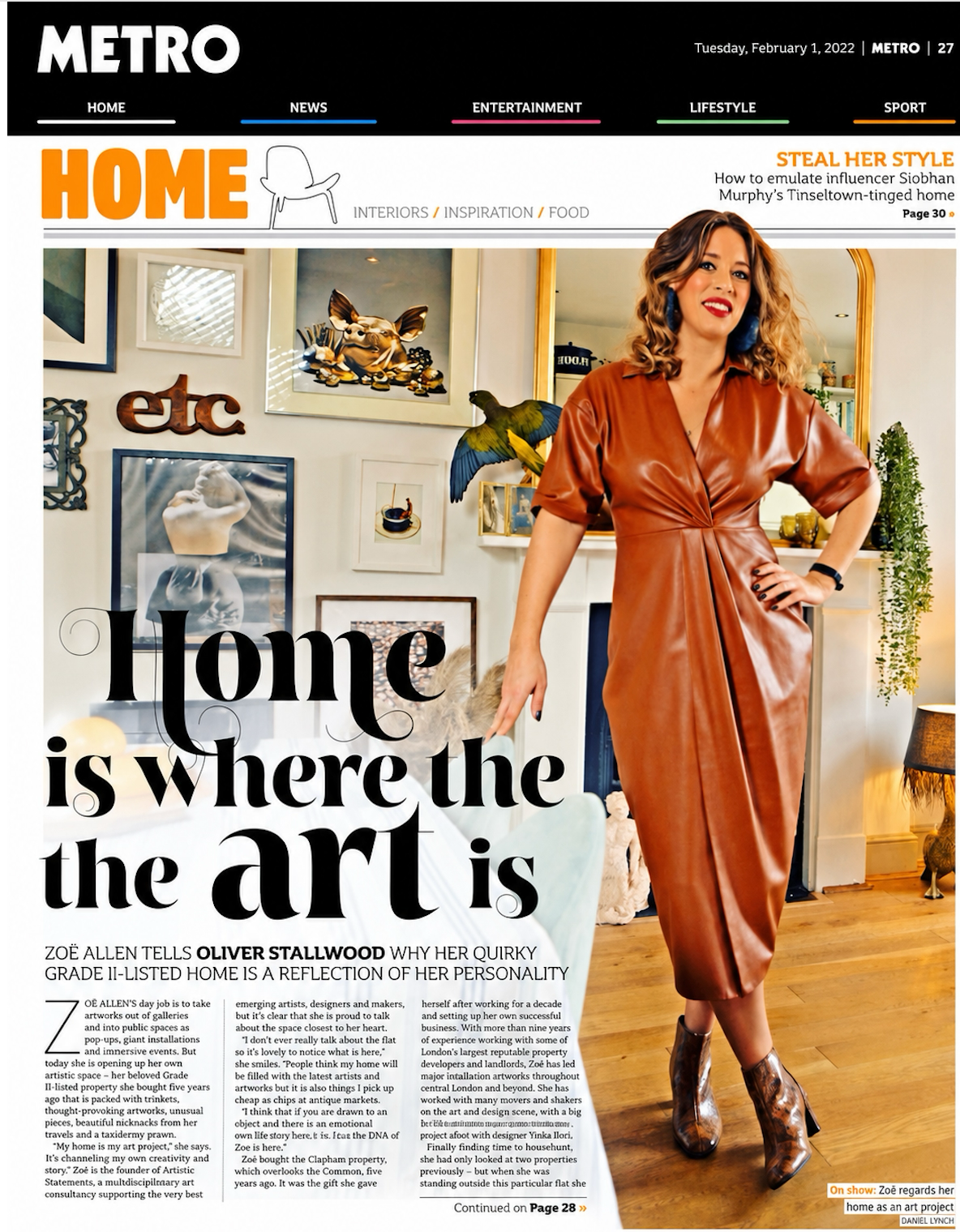 A woman with curly hair wearing a brown leather dress and brown boots stands in a decorated living room. The background features framed artwork on the wall, a parrot, and various decorative items, including plants and a lamp.
