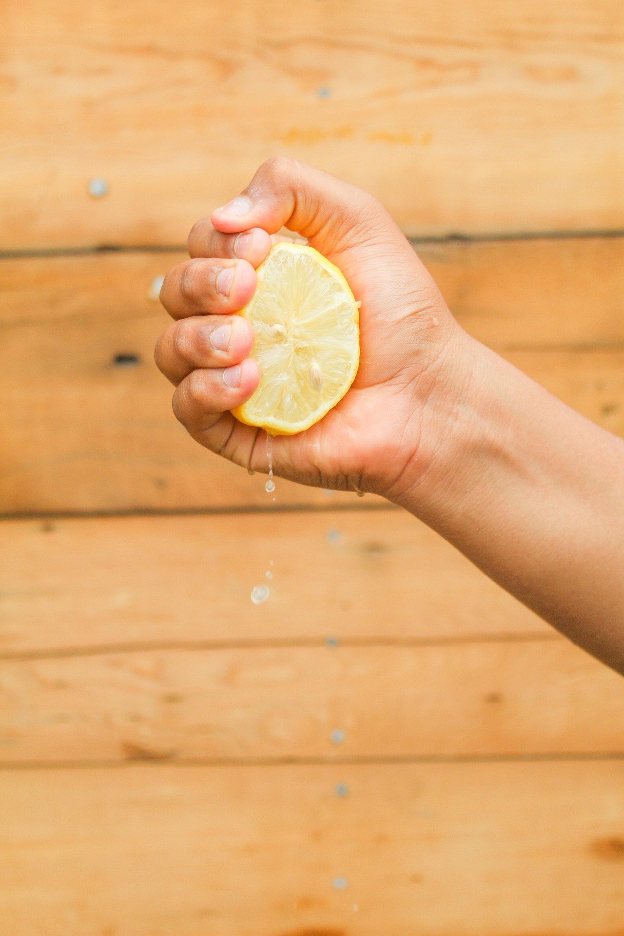 A hand squeezing a lemon, with juice droplets falling, against a wooden background.