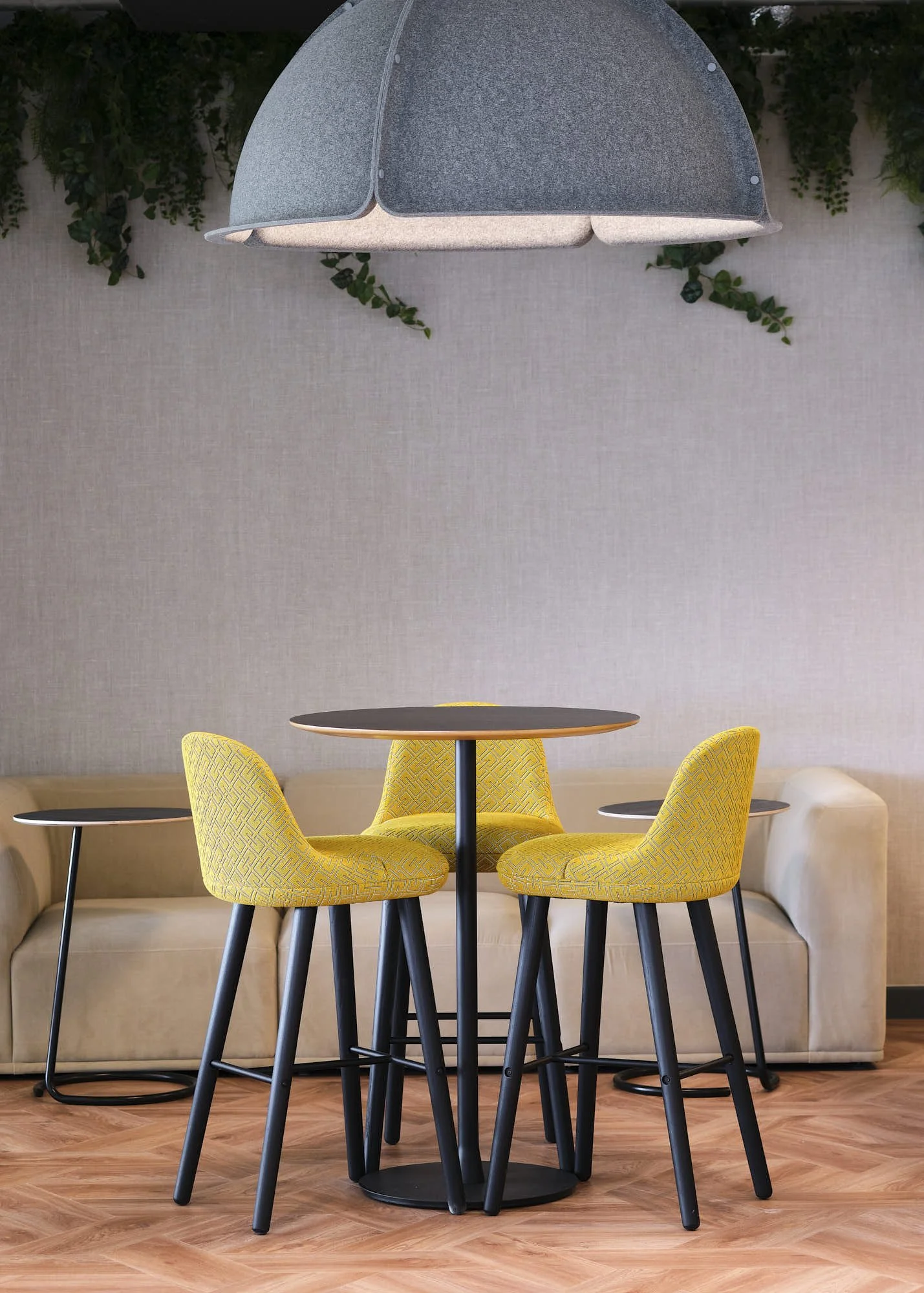 Modern dining area with a round black table surrounded by four yellow upholstered chairs with black legs, situated in front of a beige sofa, with a hanging gray pendant light and green plants on the wall above.