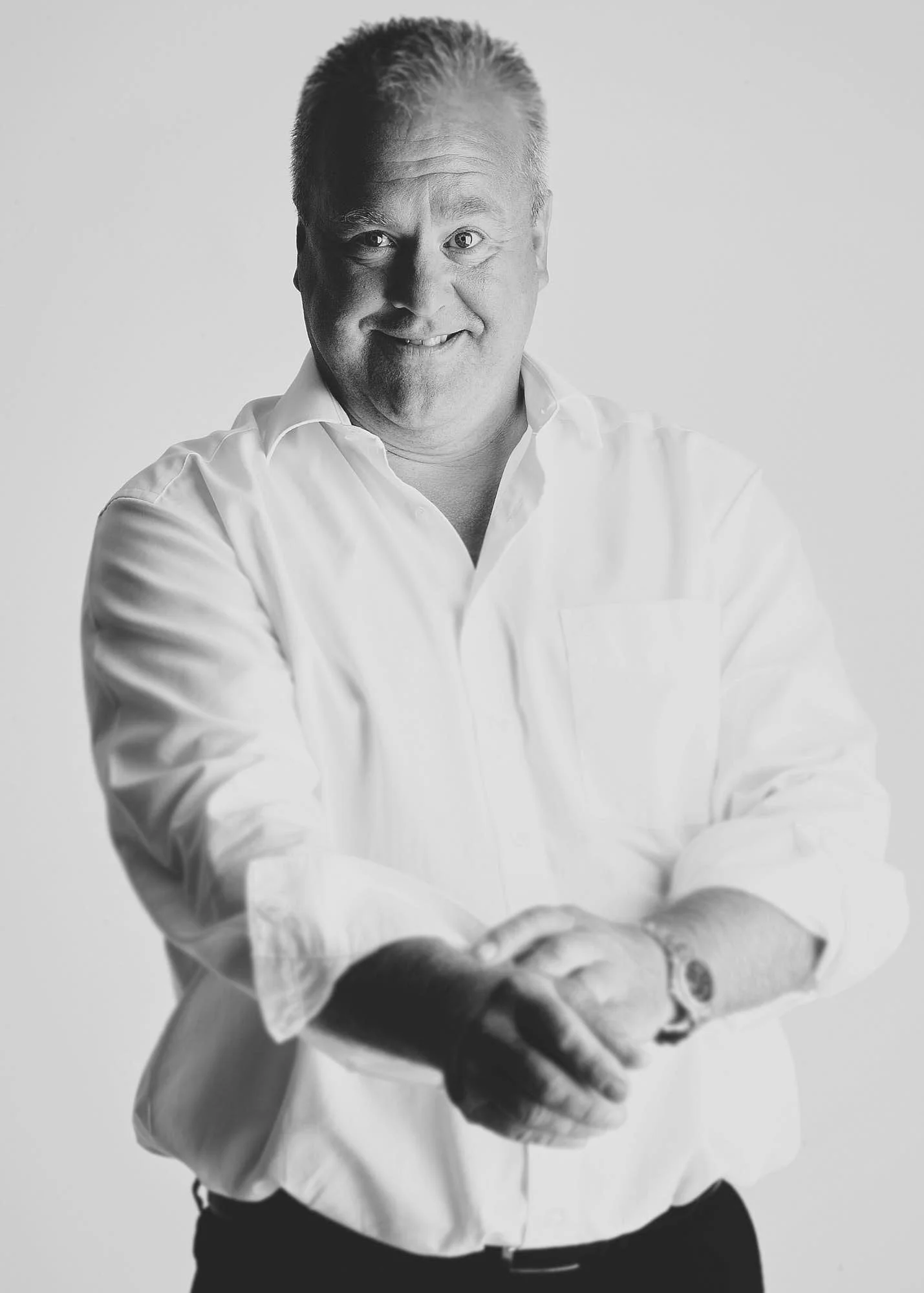 Black and white photo of a man in a white dress shirt smiling with his arms crossed and hands clasped, against a plain background.