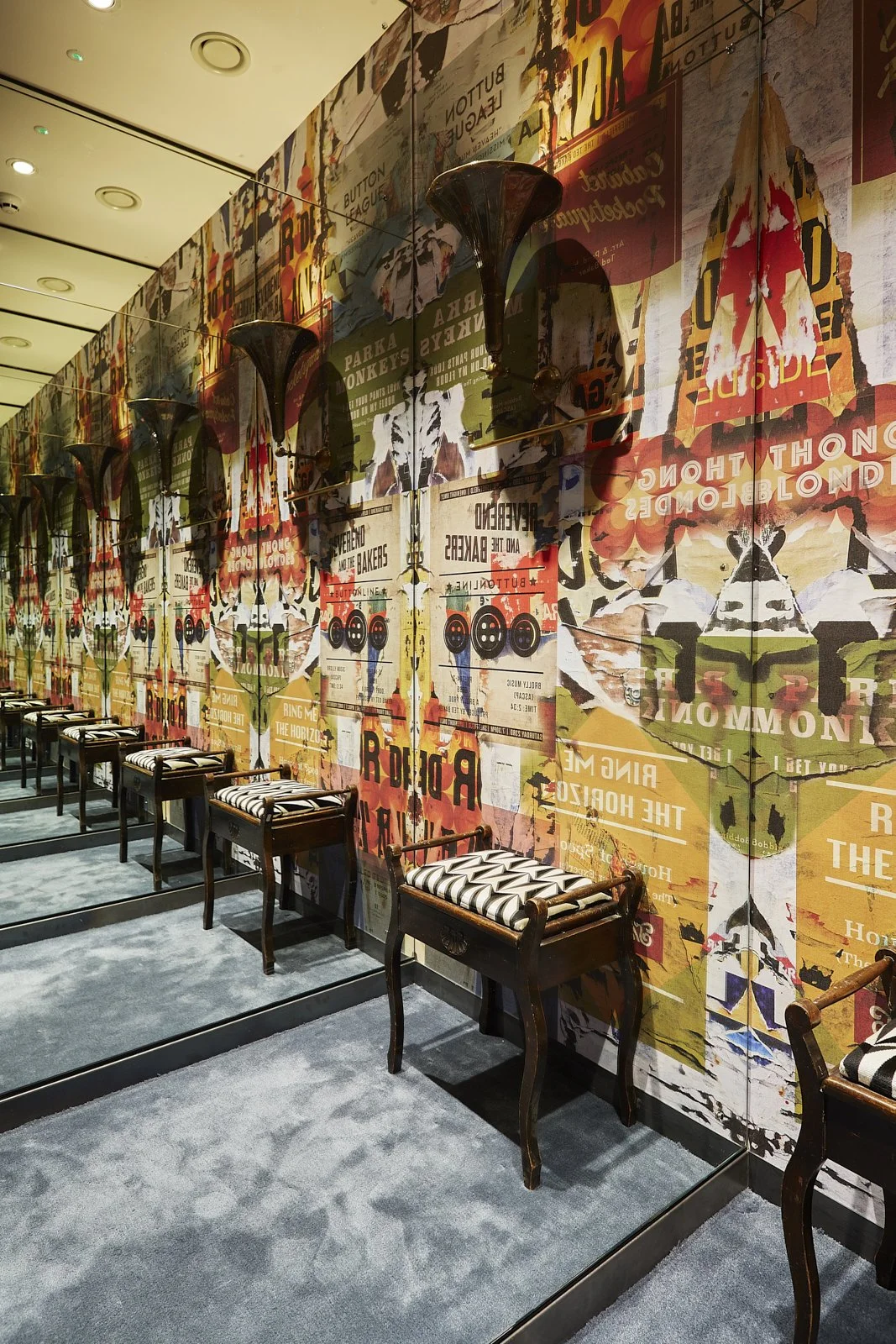 A row of vintage wooden chairs with black and white striped cushions set against a colorful, collage-style wall with posters, text, and images, reflected in a large mirror.