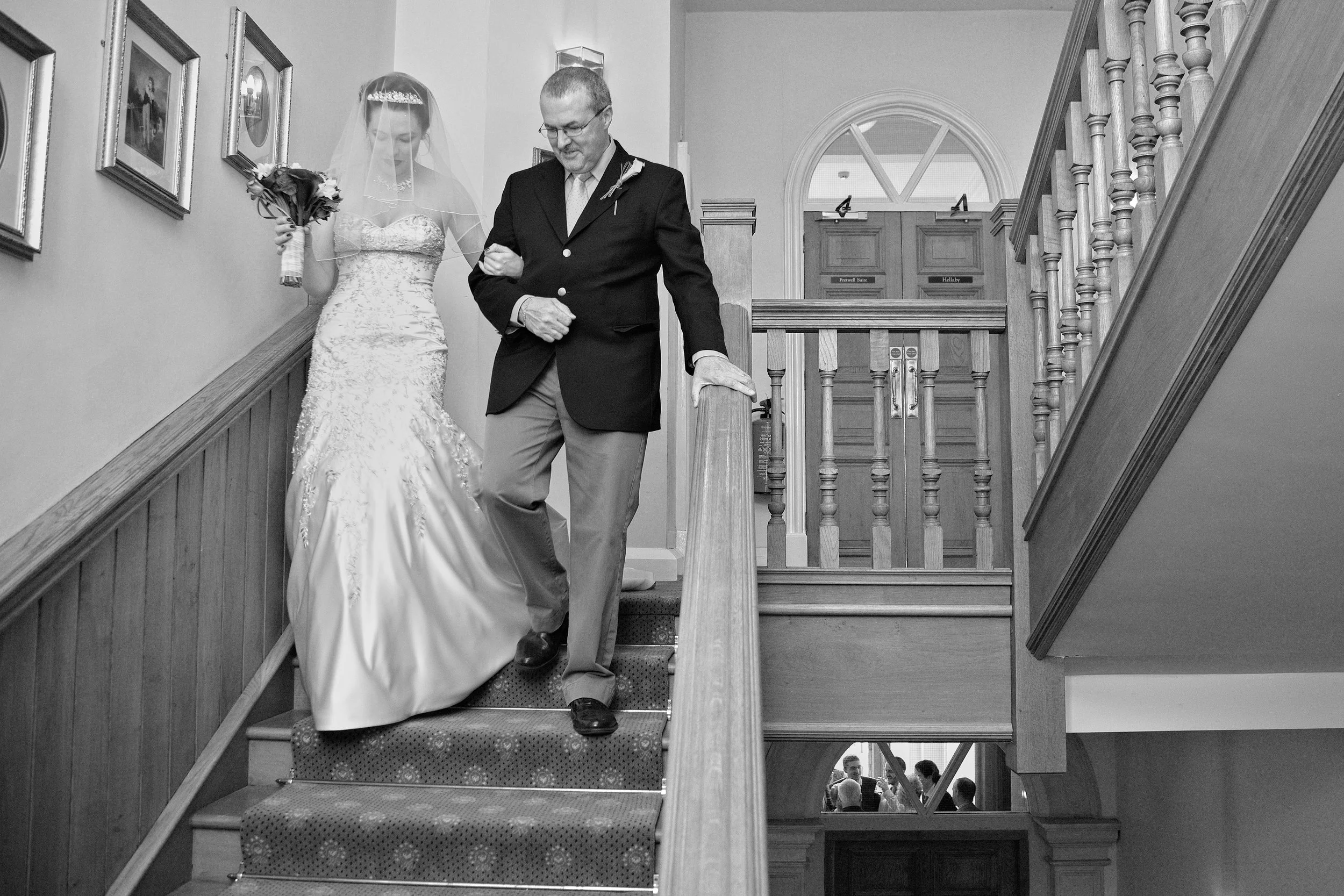 A bride in a wedding dress holding a bouquet walking down stairs escorted by a man in a suit. The staircase has wooden banisters and carpeted steps. The background shows a wooden panel with picture frames and a door.