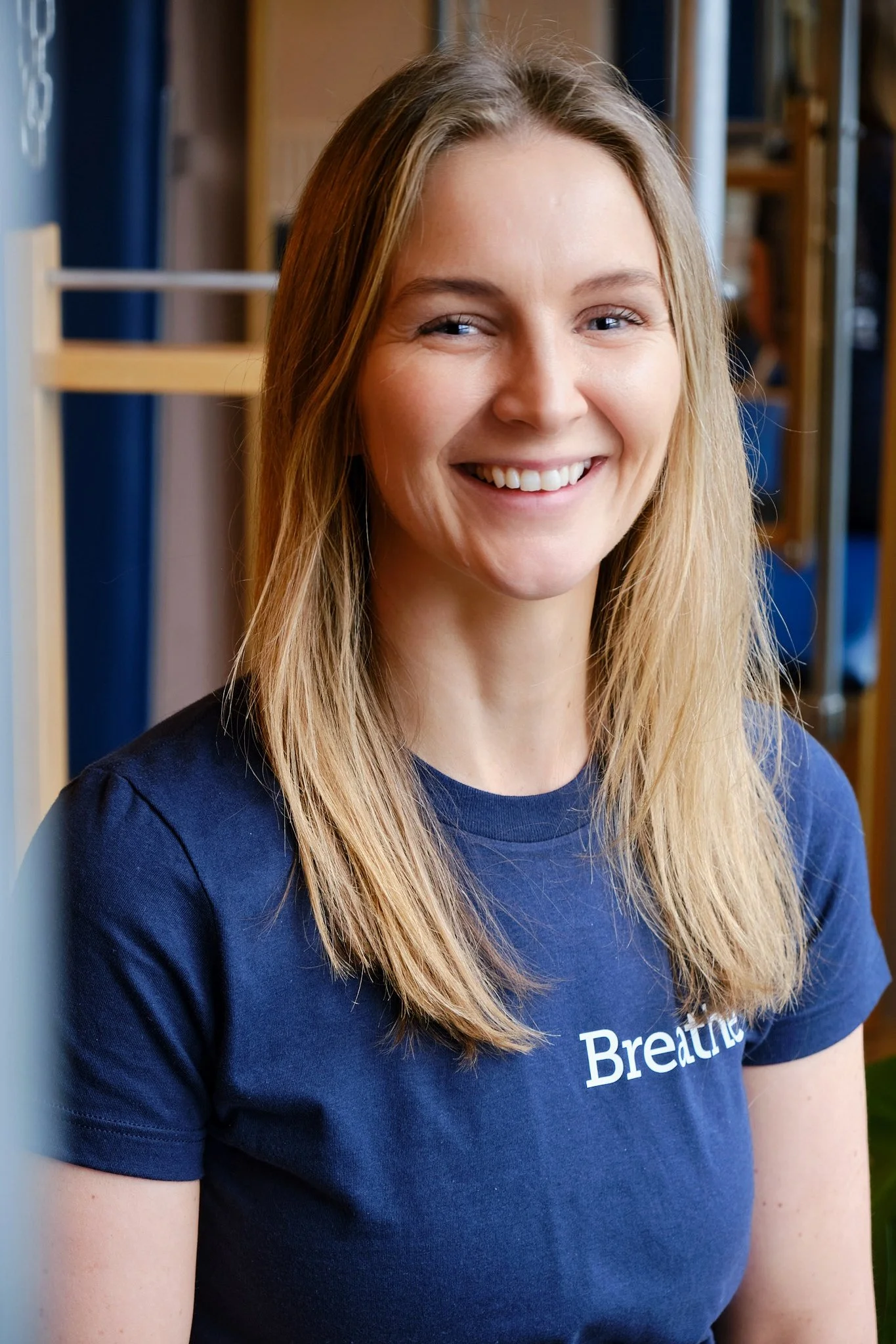 A young woman with long blonde hair smiling, wearing a blue T-shirt that says 'Breathe' on it, with a background of wooden shelves and some fabric.