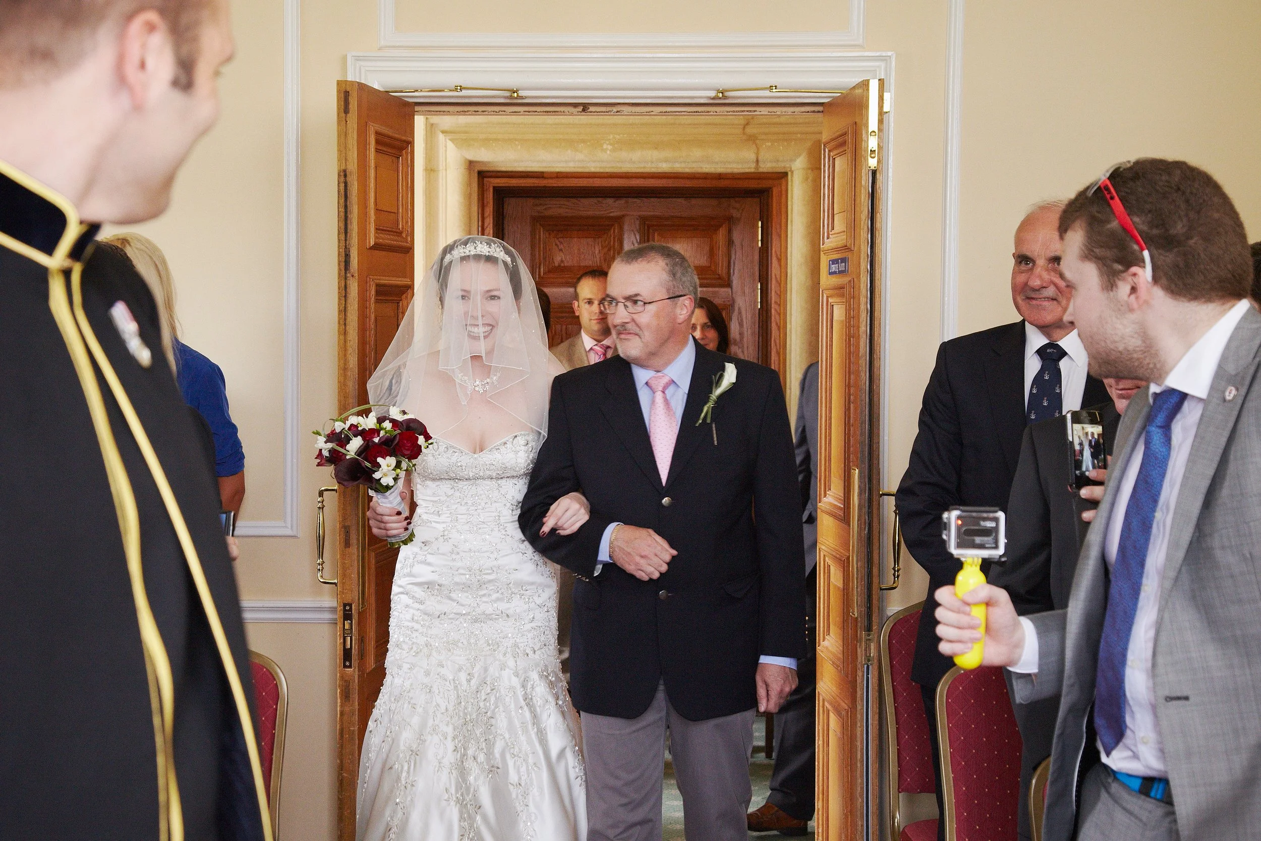 A bride in a white wedding dress with lace details holding a bouquet of red and white flowers, walking down the aisle with a man in a black suit. The scene is inside a room with wooden double doors in the background. Several people are present, inclu