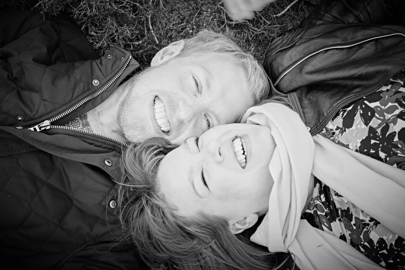 Two people, a man and a woman, lying on the ground with their heads close together, smiling and appearing happy.