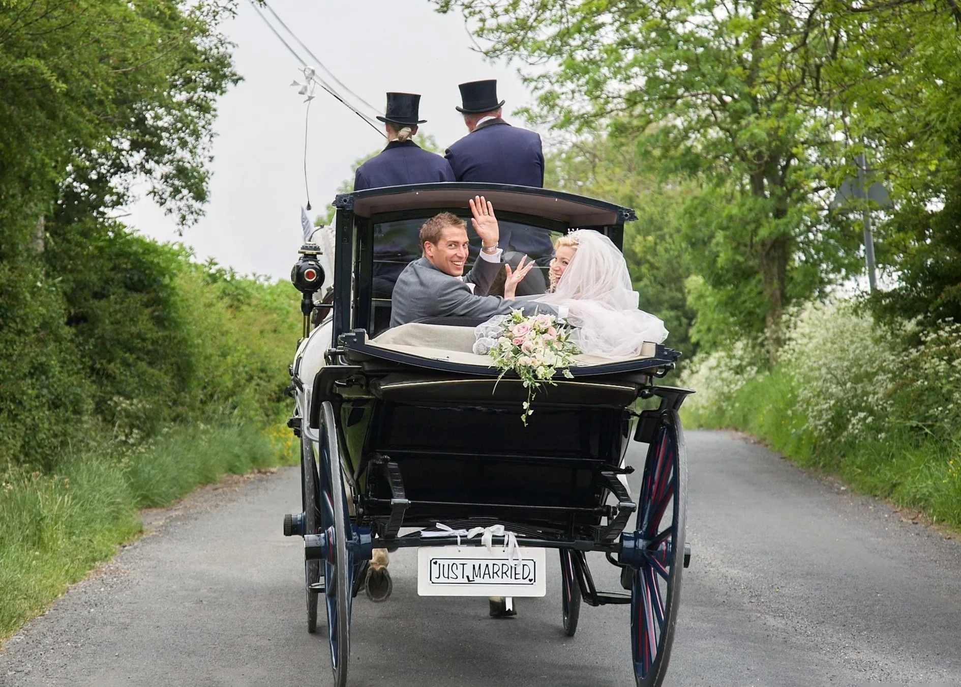 A newlywed couple riding in a horse-drawn carriage with a sign that reads 'Just Married,' waving and smiling, surrounded by green trees on a rural road.