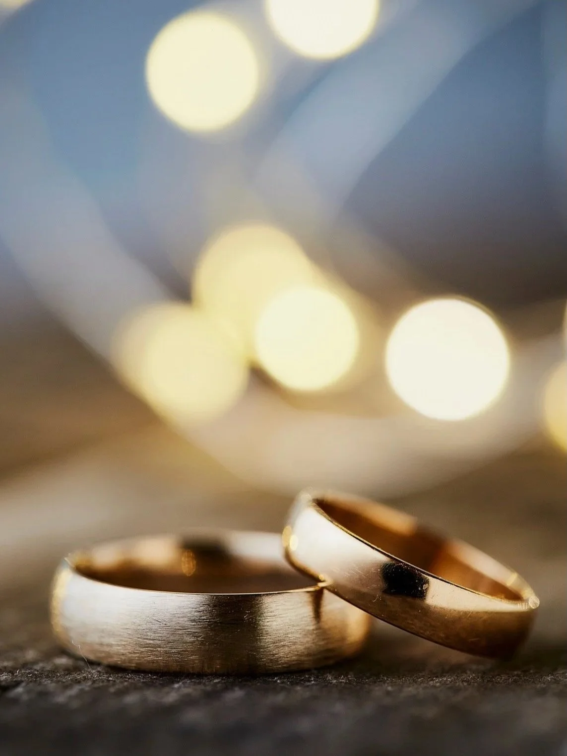 Close-up of two gold wedding bands on a dark surface, with blurred yellow and white lights in the background.