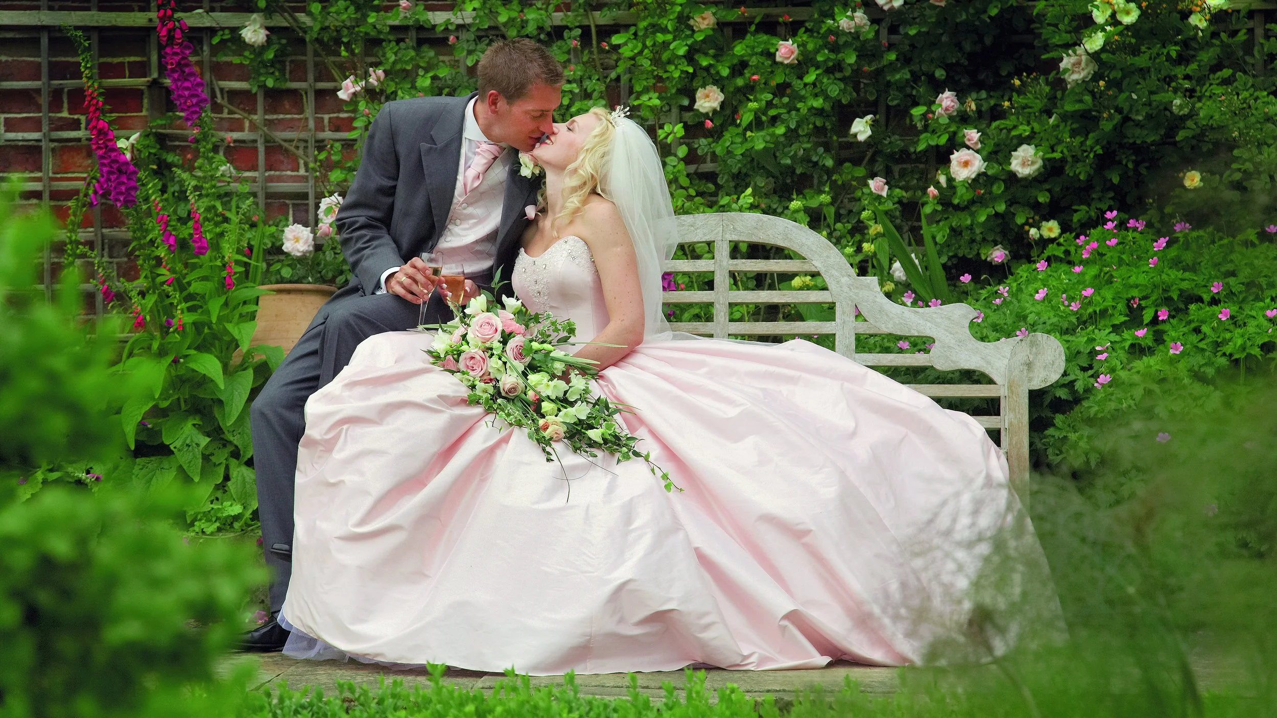 A newlywed couple sharing a kiss while sitting on a garden bench surrounded by flowers.
