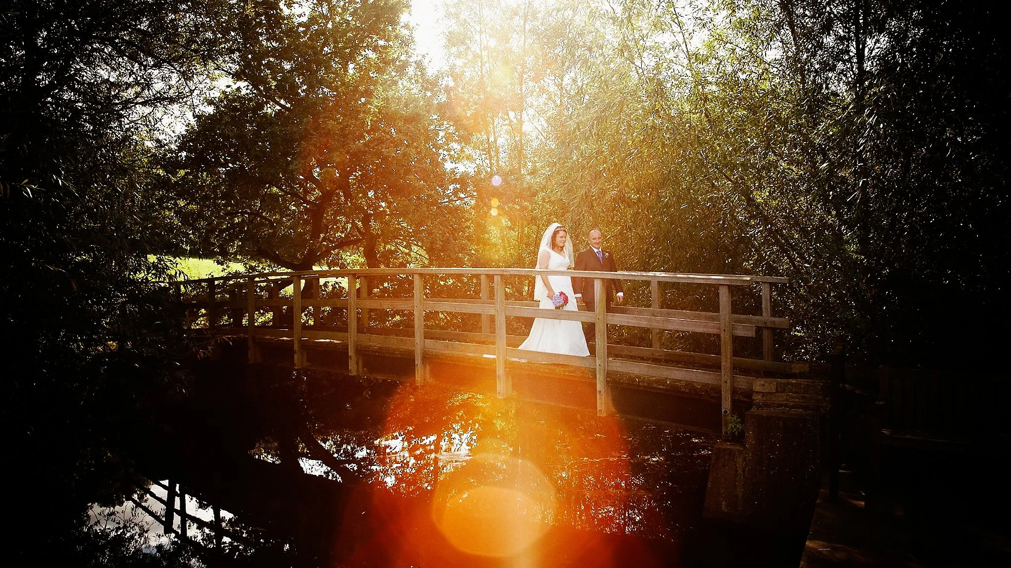 A bride and groom walking on a wooden bridge surrounded by trees with sunlight shining through.