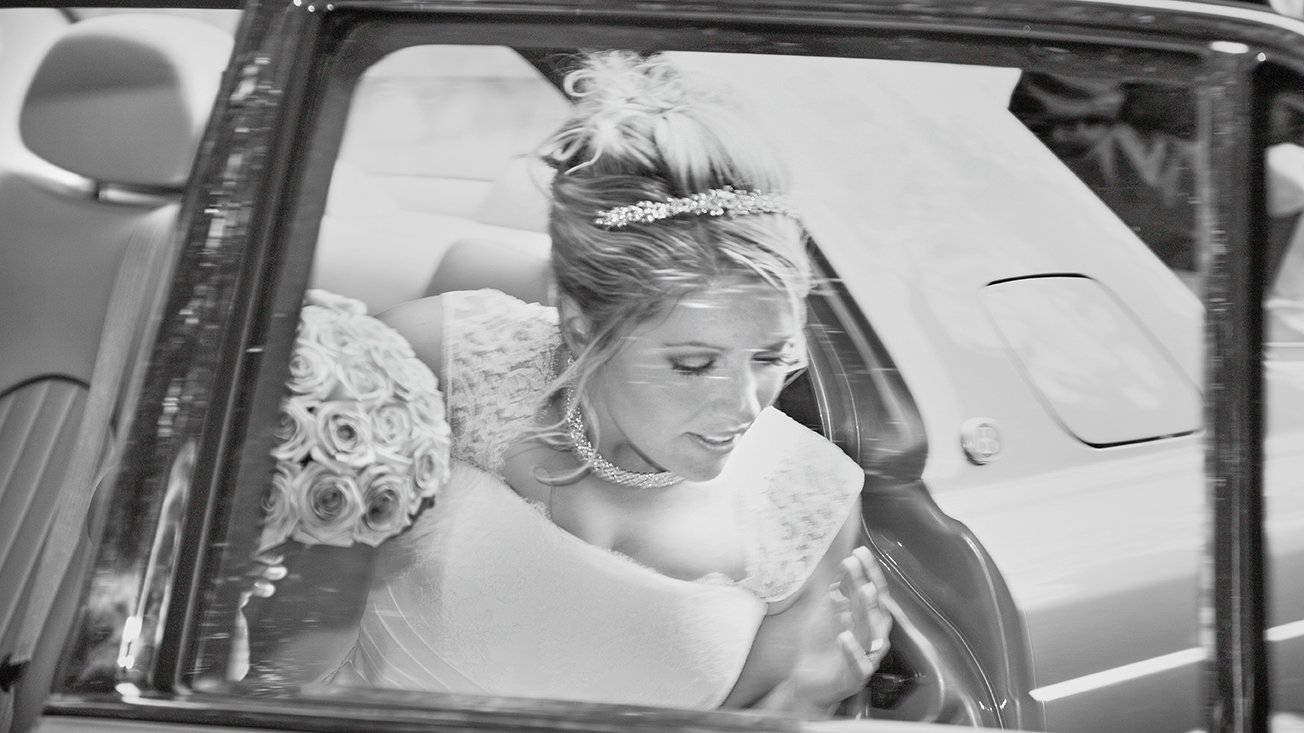 A bride in a wedding dress with a bouquet of roses and a tiara on her head, sitting in a car, looking down with her hands pressed together.