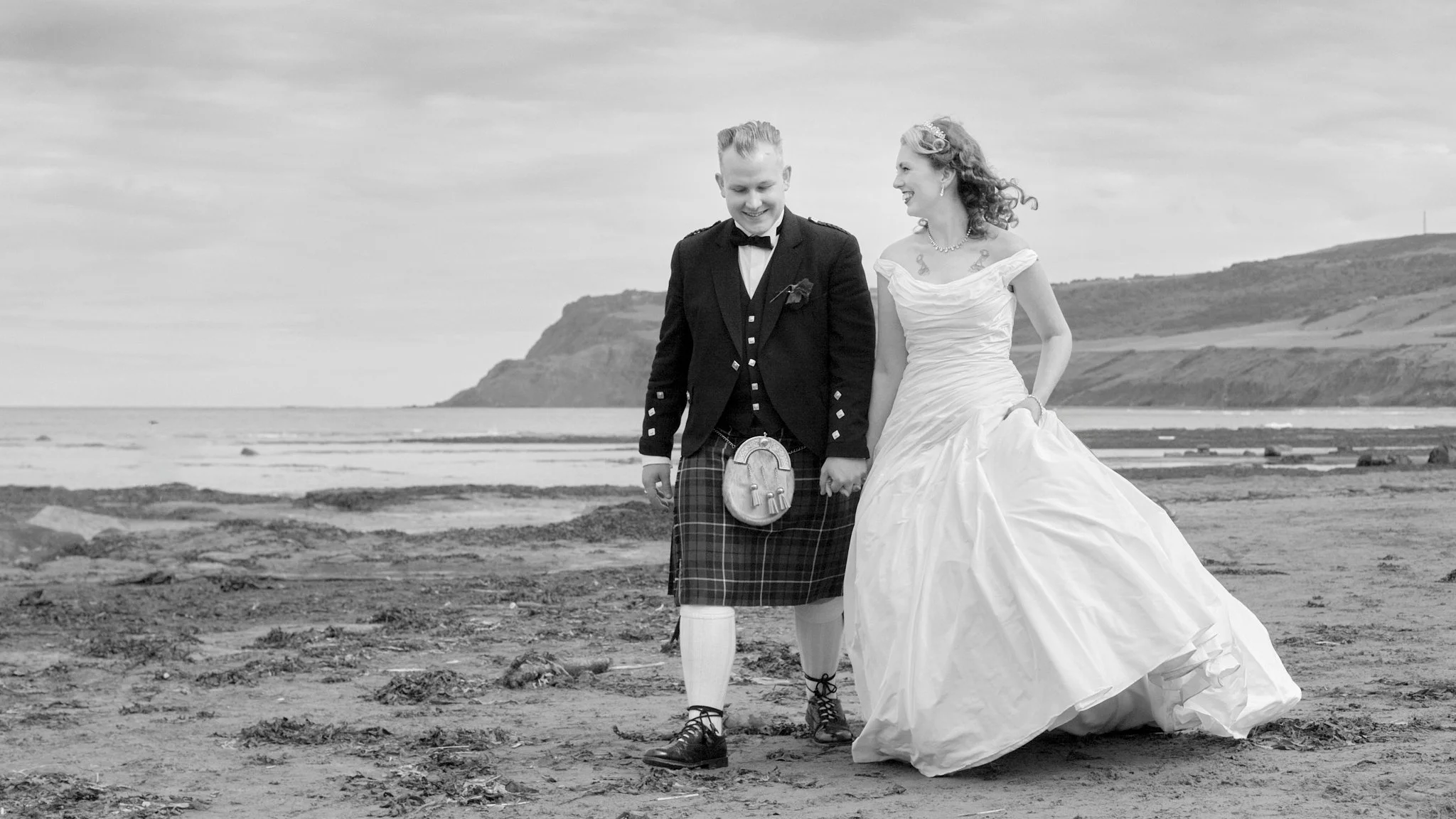 A black and white photo of a wedding couple walking on a beach, holding hands and smiling. The groom wears traditional Scottish attire, and the bride wears a wedding dress with off-the-shoulder straps.