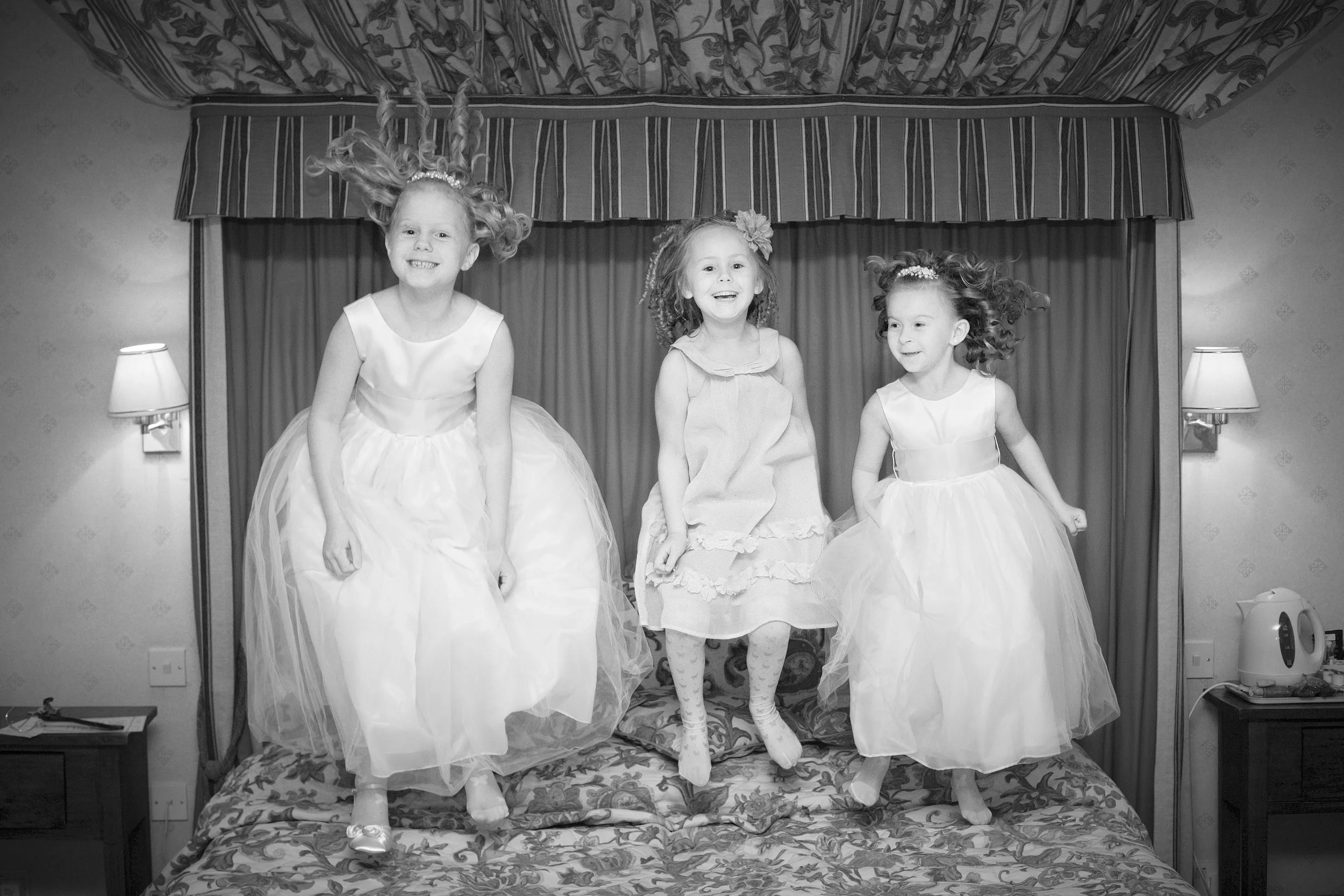 Black and white photo of three young girls standing on a bed with a floral bedspread and headboard, smiling and holding hands, with a curtain and lamps on either side in the background.