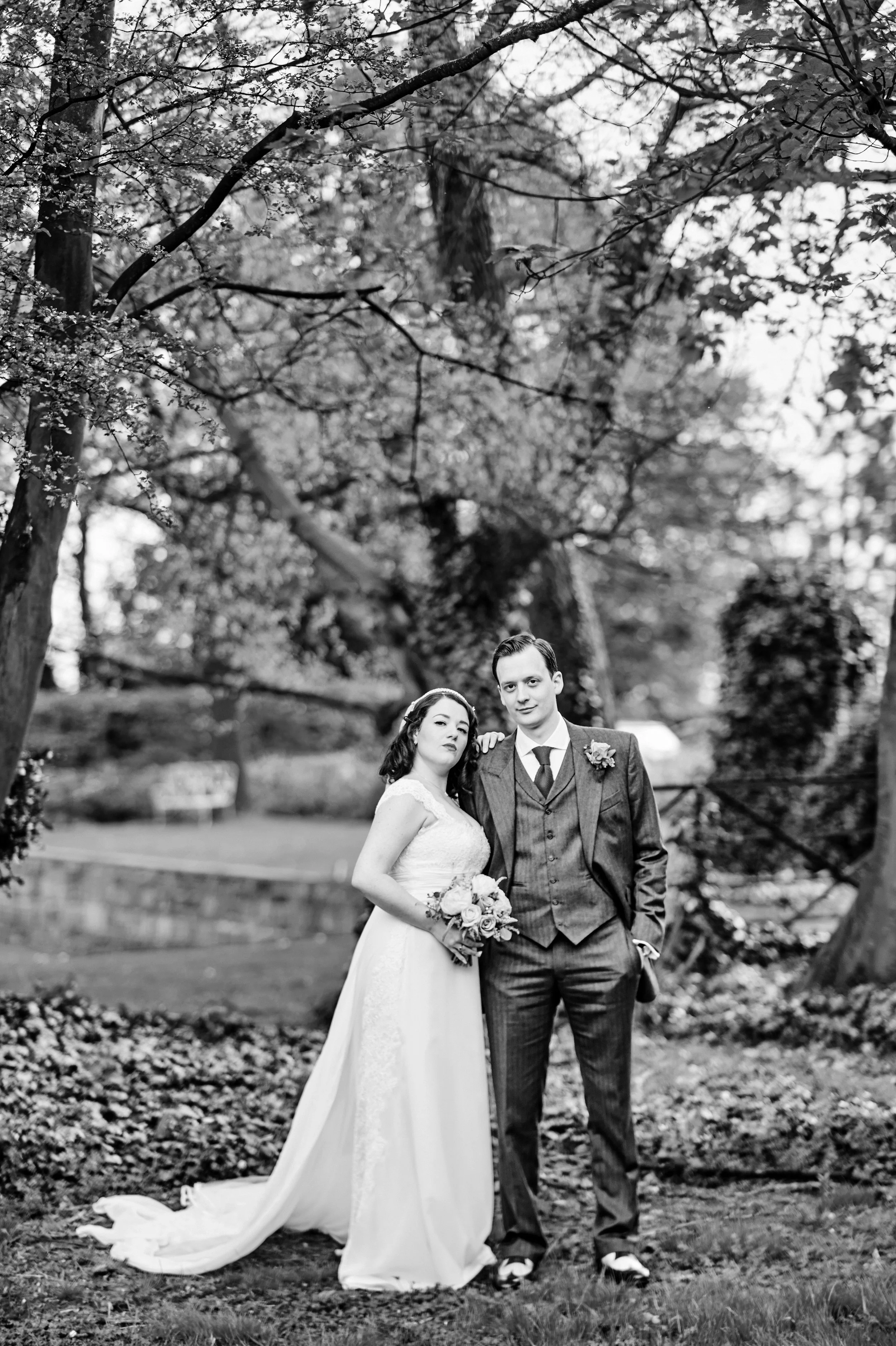 Black and white photo of a bride and groom standing outdoors in a park-like setting with trees and a wooden fence in the background. The bride wears a long wedding dress and holds a bouquet, and the groom wears a suit with a tie and boutonnière. They