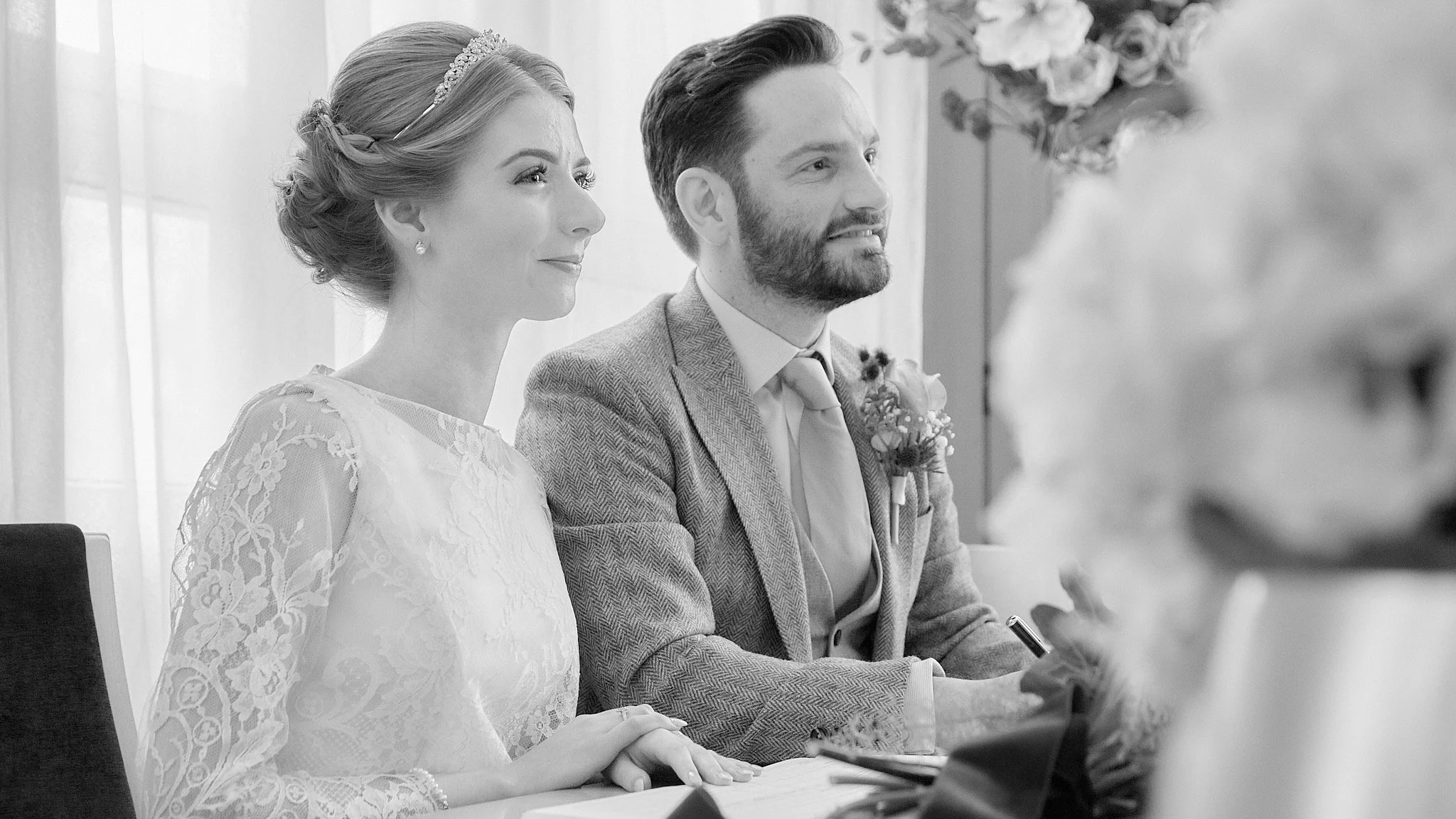 A black-and-white photo of a bride and groom at a wedding ceremony, sitting side by side and listening to a person speaking in front of them.