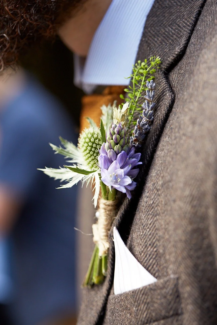 Close-up of a boutonniere with purple, white, and green flowers pinned to a man's brown suit jacket at a formal event.