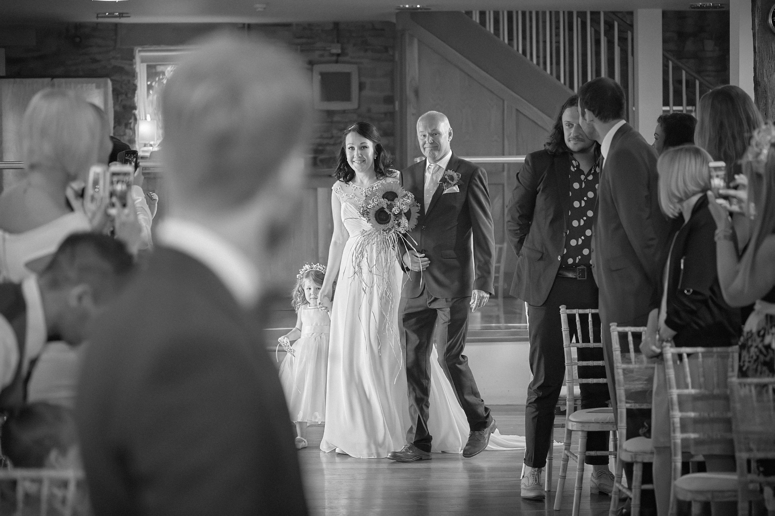 In black and white, a bride walks down the aisle holding a bouquet of flowers, accompanied by a man in a suit. Guests are seated and taking photos of the moment in a rustic wooden venue.