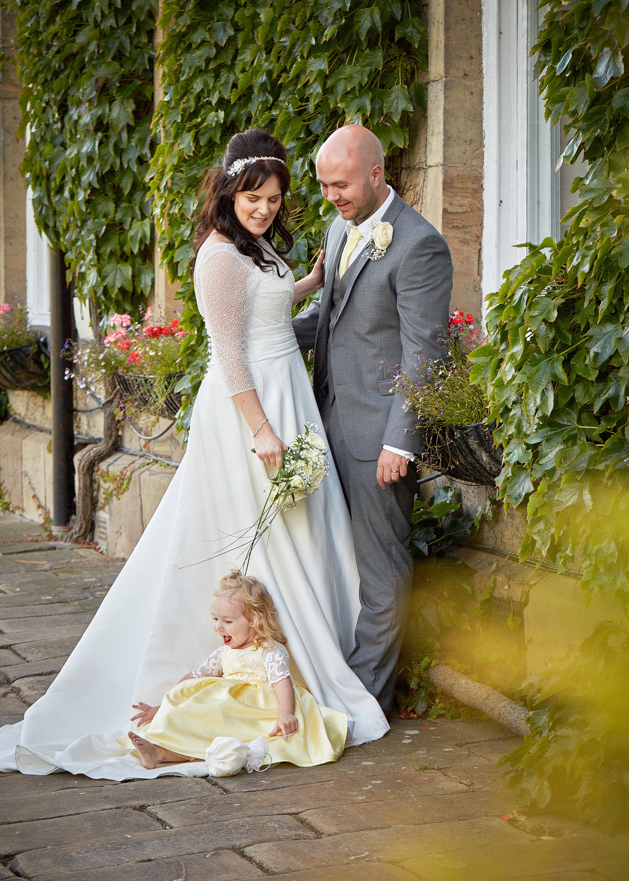 A bride and groom are standing outdoors by a plant-covered wall. The bride is wearing a white wedding dress and a floral headpiece, holding a bouquet of white flowers. The groom is dressed in a gray suit with a light yellow tie and boutonniere. Two y
