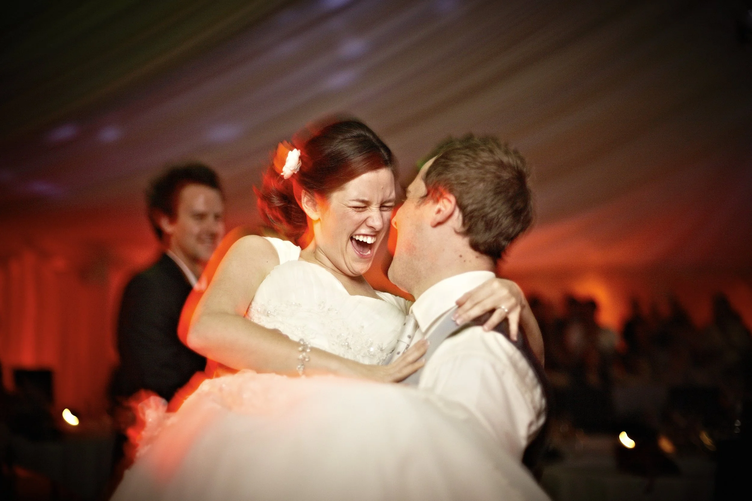 A bride and groom dancing and smiling during their wedding reception, with other guests in the background.