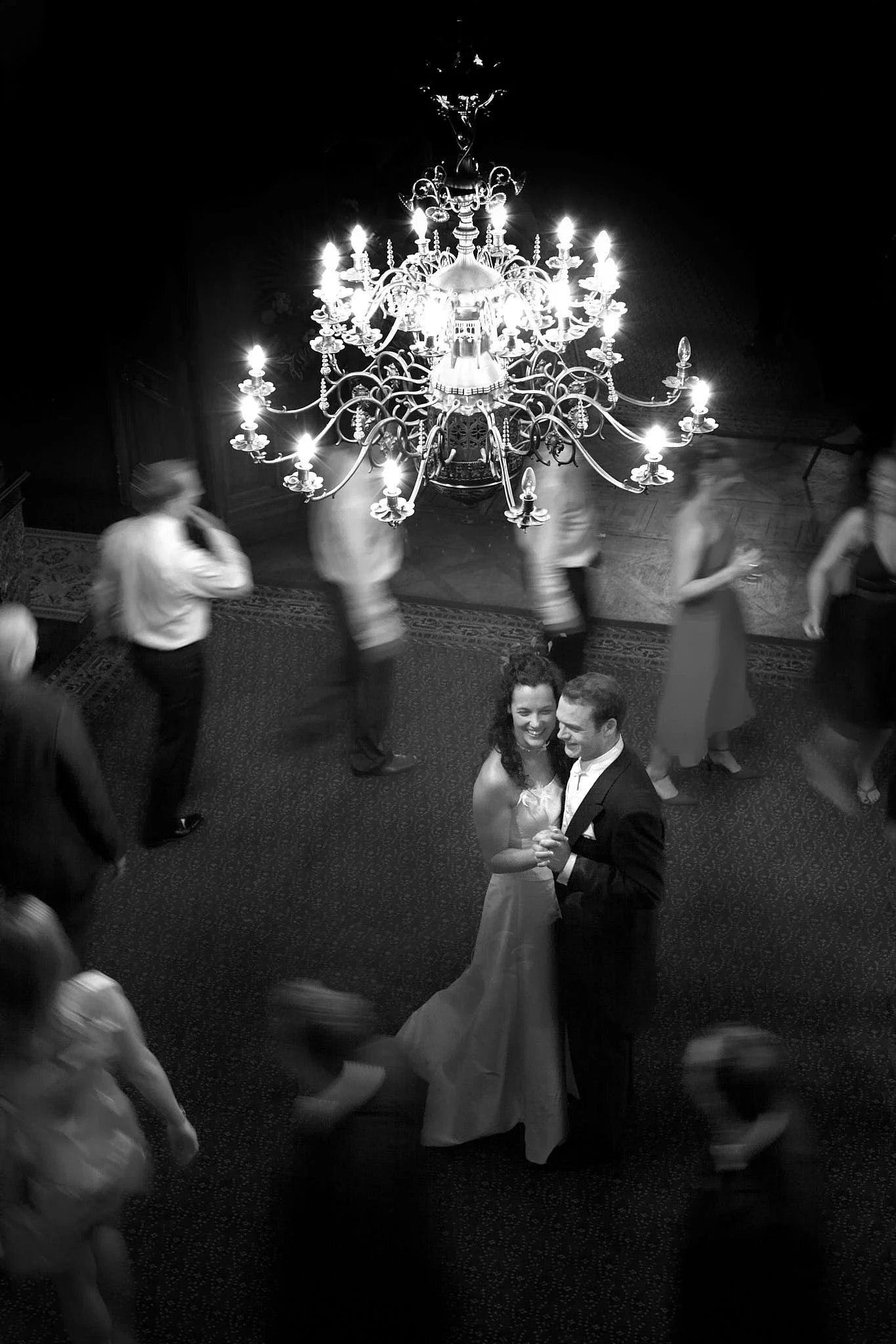 A black and white photo of a couple dancing closely in a grand hall with a large chandelier overhead and people dancing around them.