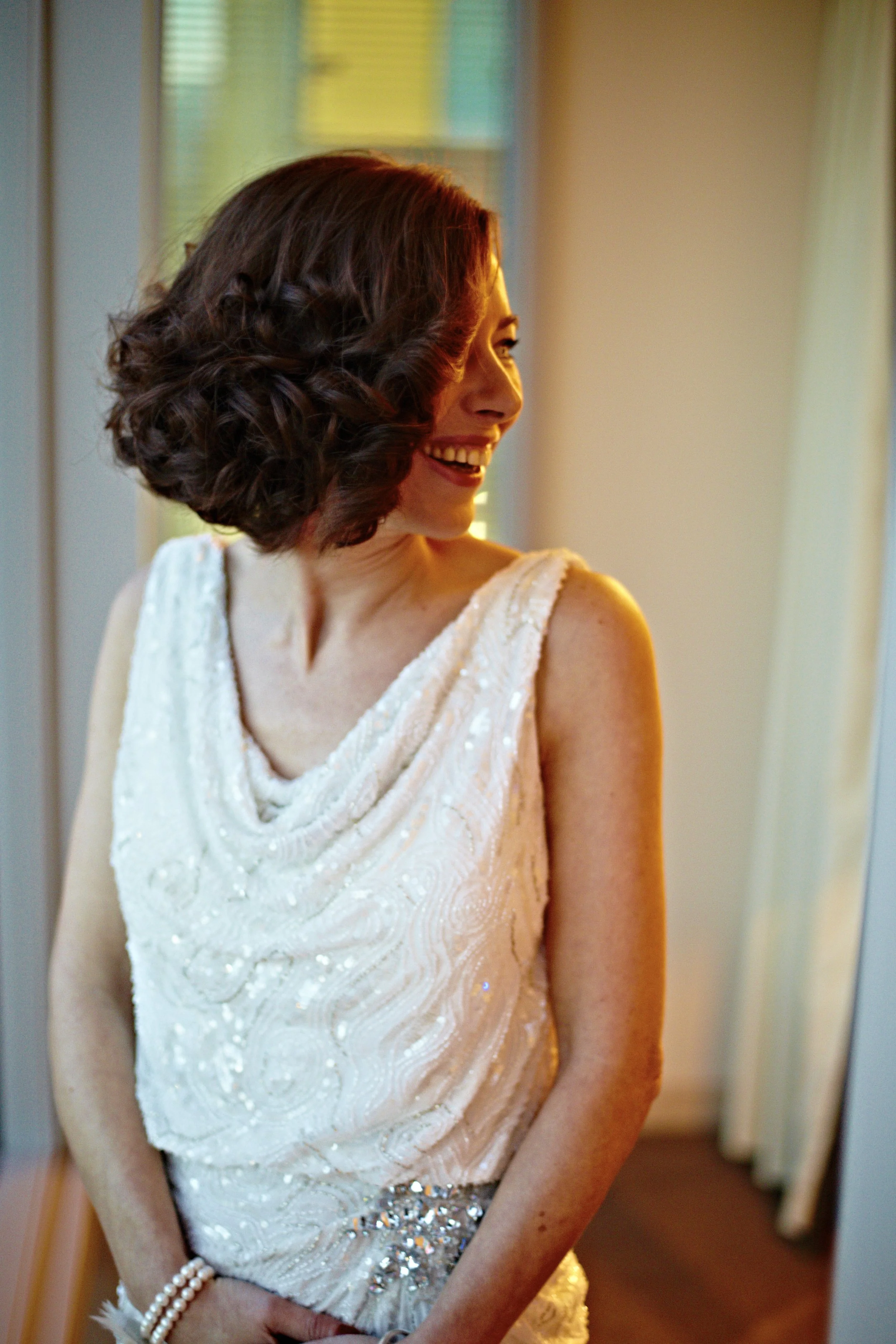 A woman with short, wavy brown hair in a white embellished dress, smiling and looking to her left, in a softly lit room with green and yellow curtains in the background.