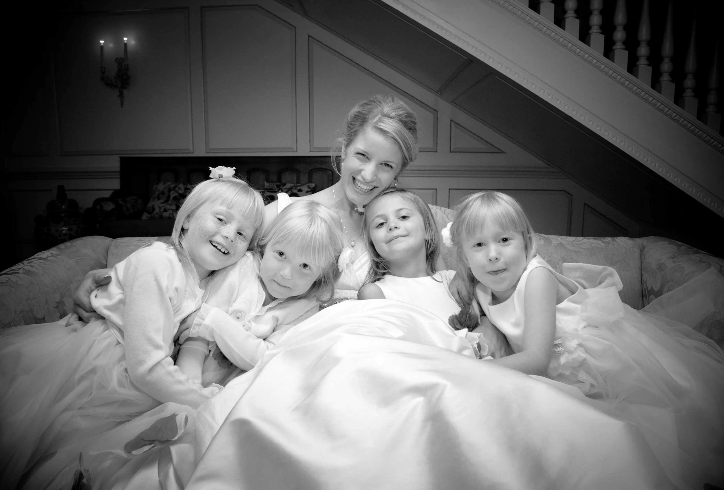 A black and white photo of a woman and five young girls sitting closely together on a bed, smiling and posing for the camera in a cozy, decorated room.