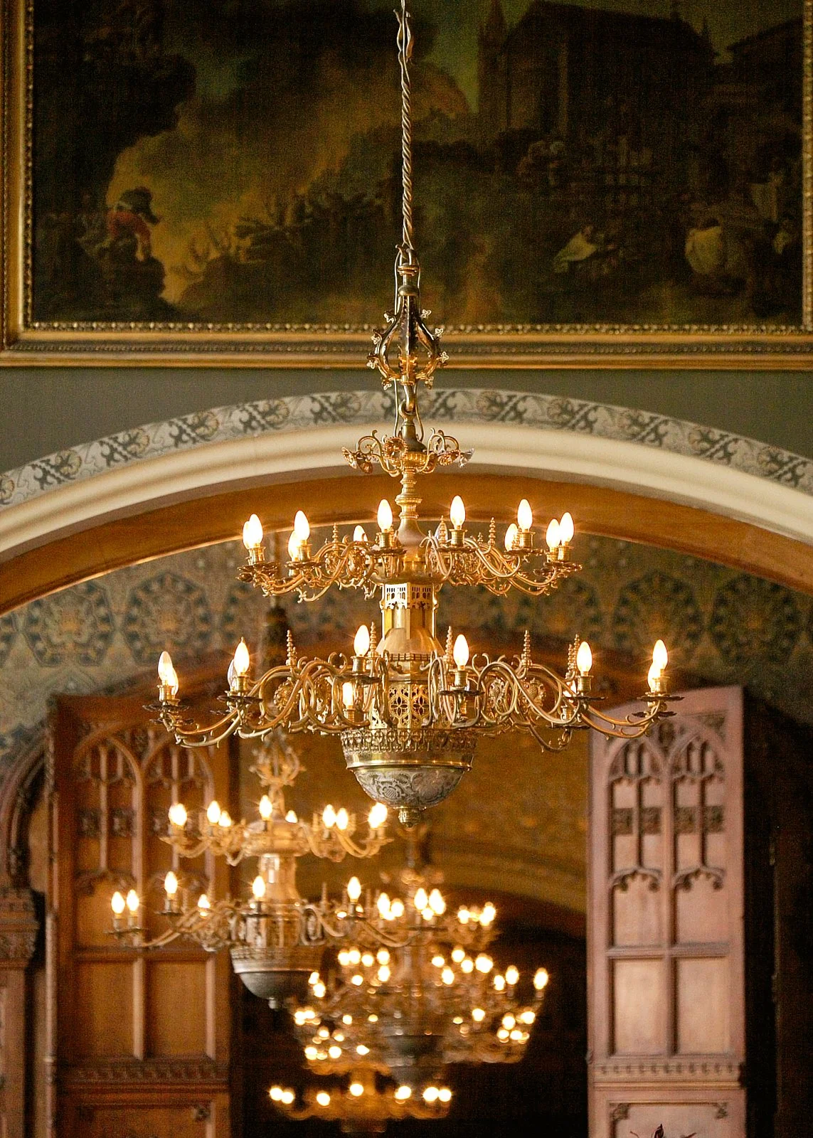 Multiple ornate gold chandeliers hanging from a ceiling in an elegant interior, with wooden doors and decorative arches, illuminated by candle-like light bulbs.
